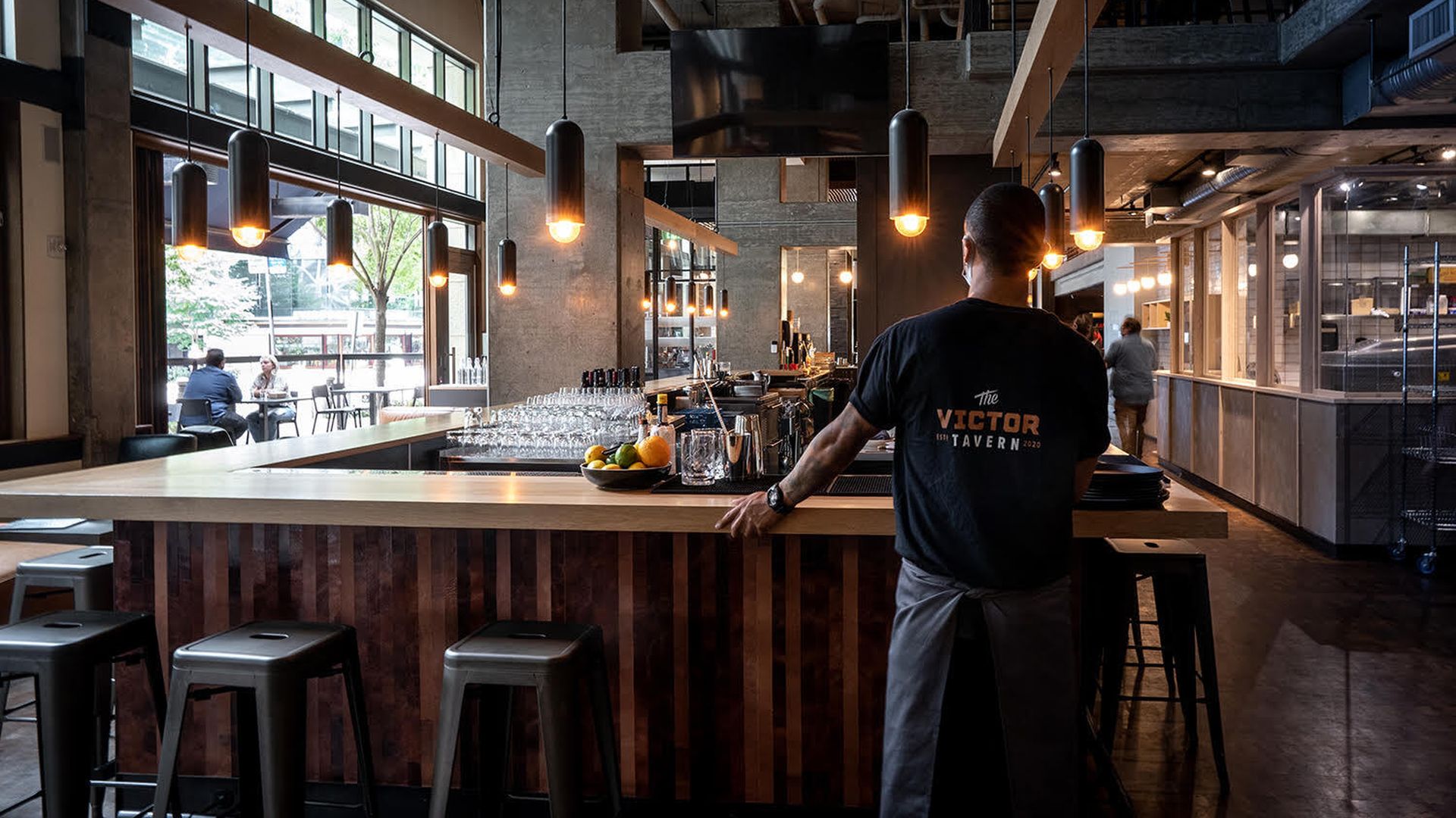 A restaurant worker stands on the outer edge of an empty barstool seating area facing a kitchen.