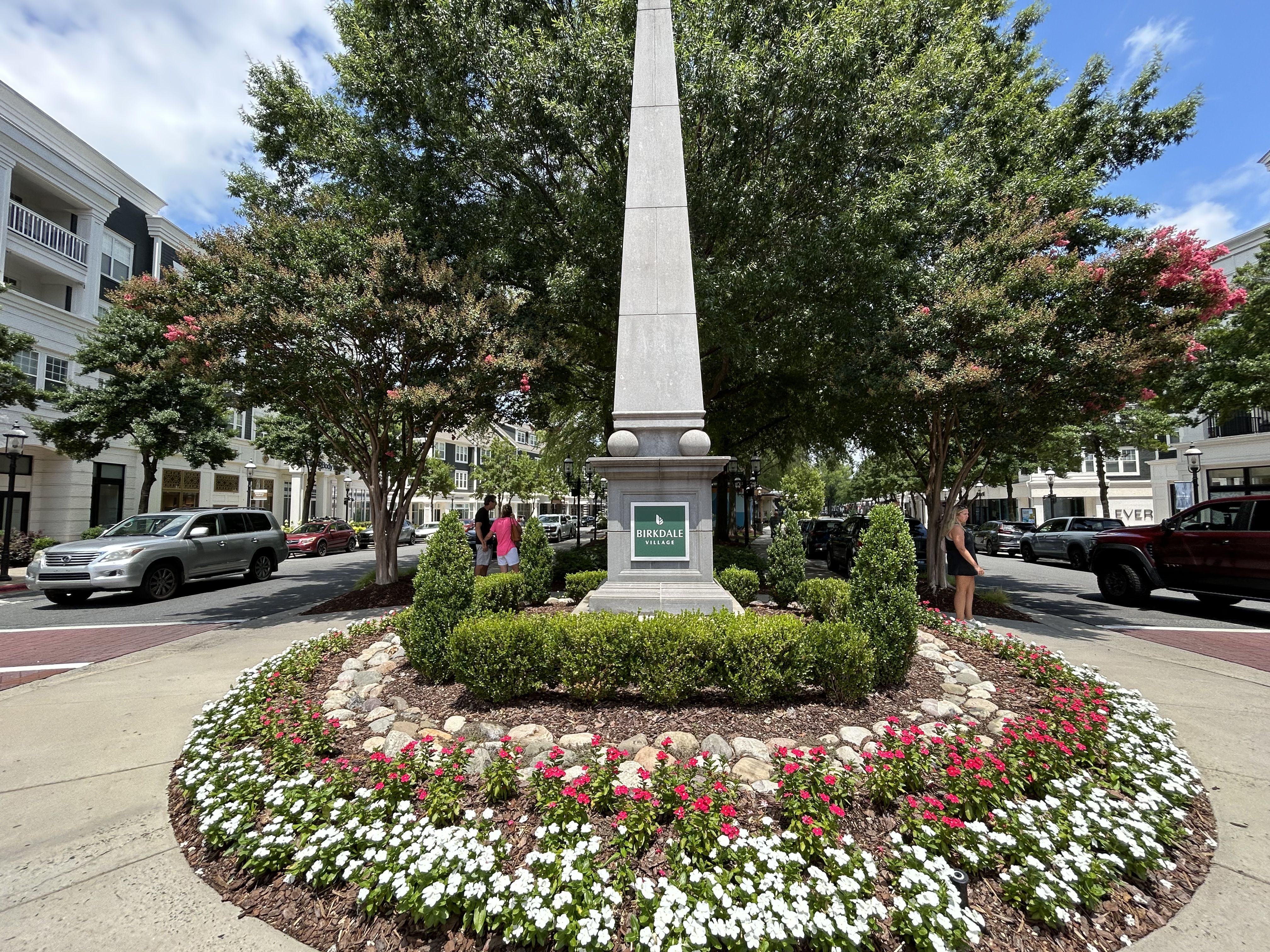 Obelisk monument with a green "Birkdale Village" sign surrounded by manicured bushes and red and white flowers in a town street with trees and parked cars under a partly cloudy sky.