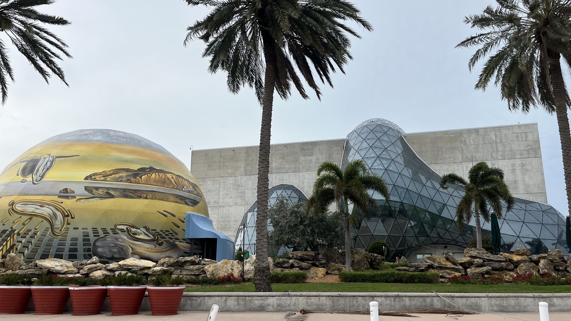 Wide view of tall palm trees framing a modern campus or park with geometric glass domes and a large yellow sphere featuring automotive imagery; stone wall, red planters, and pale blue sky.