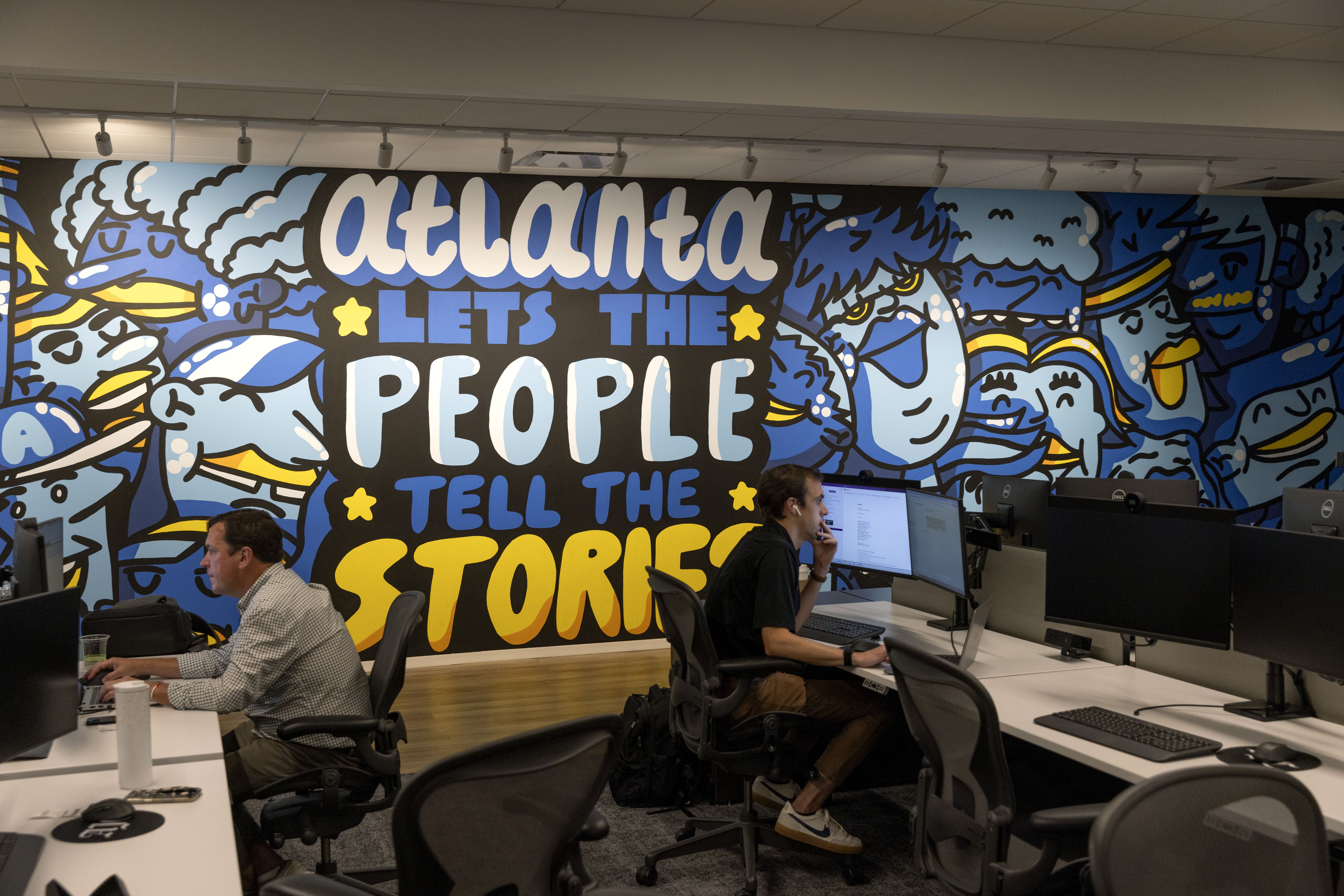 Office with two men working at desks in front of a large colorful mural that reads "Atlanta lets the people tell the stories" with blue and yellow cartoon faces on a black background.
