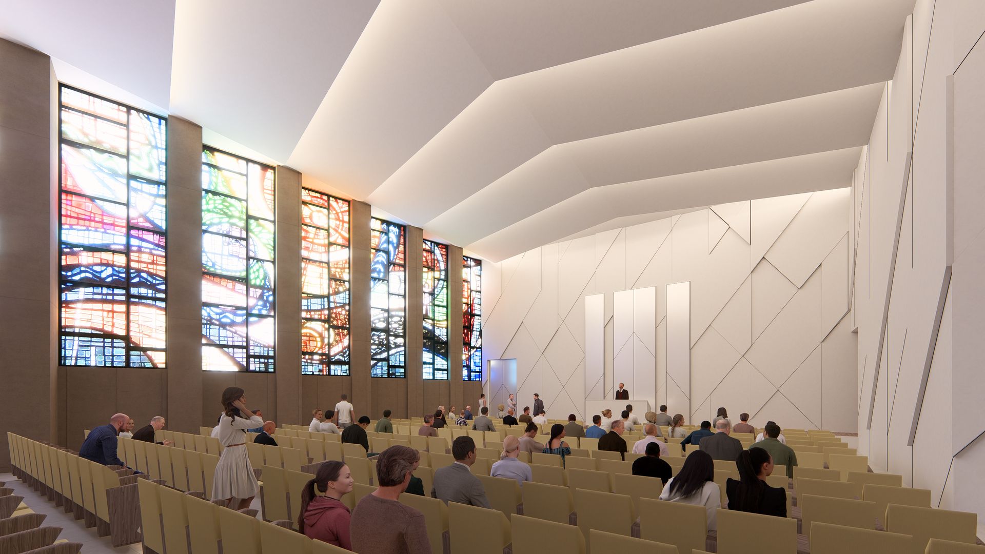Interior of a modern chapel with beige seats, geometric white walls, and large colorful stained-glass windows on the left. People are seated facing a speaker at a white podium.