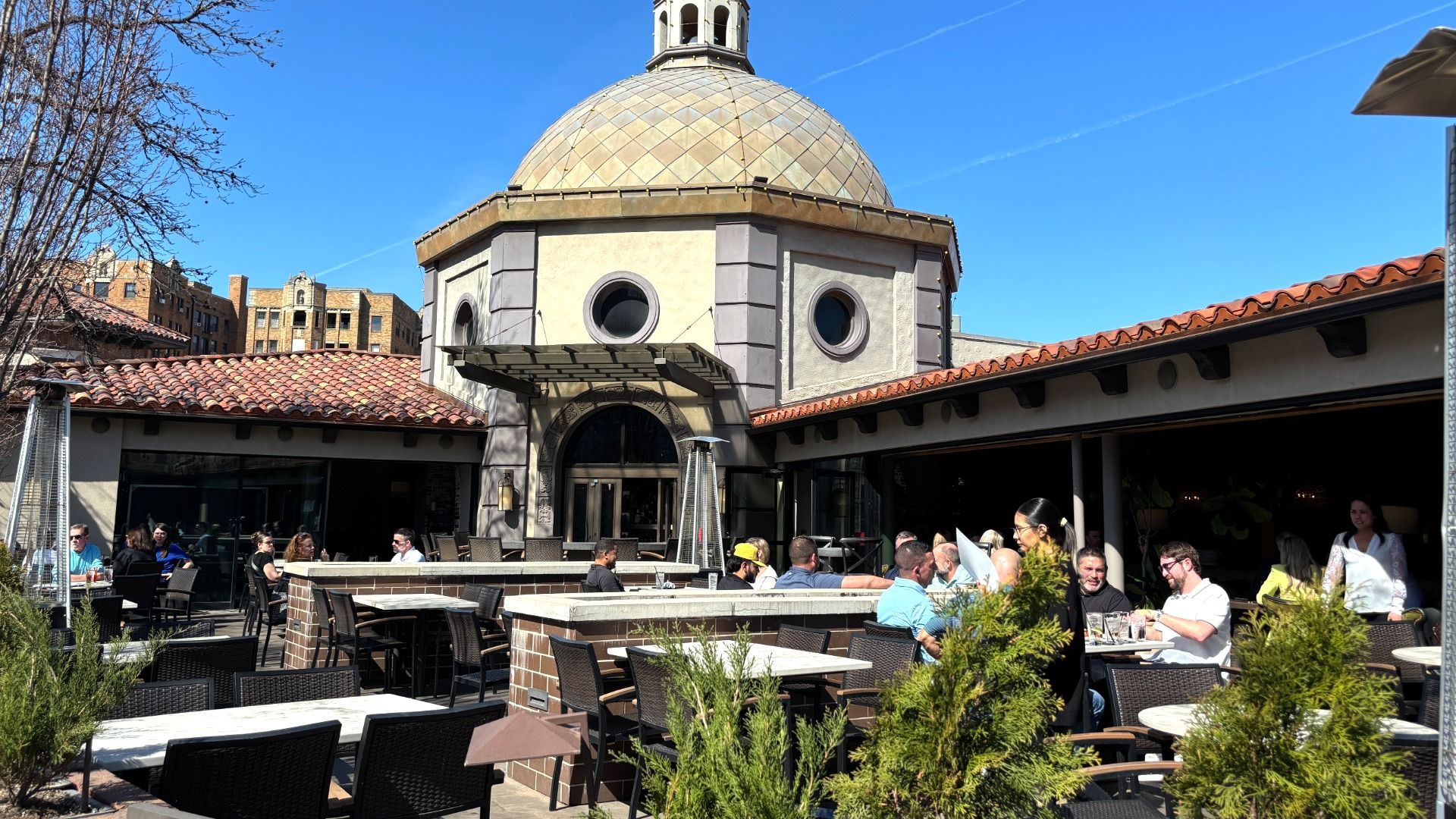 Outdoor restaurant patio around a domed building with circular windows and a tiled dome. Guests sit at tables beneath a red-tiled roof, with a bright blue sky and city buildings in the background.