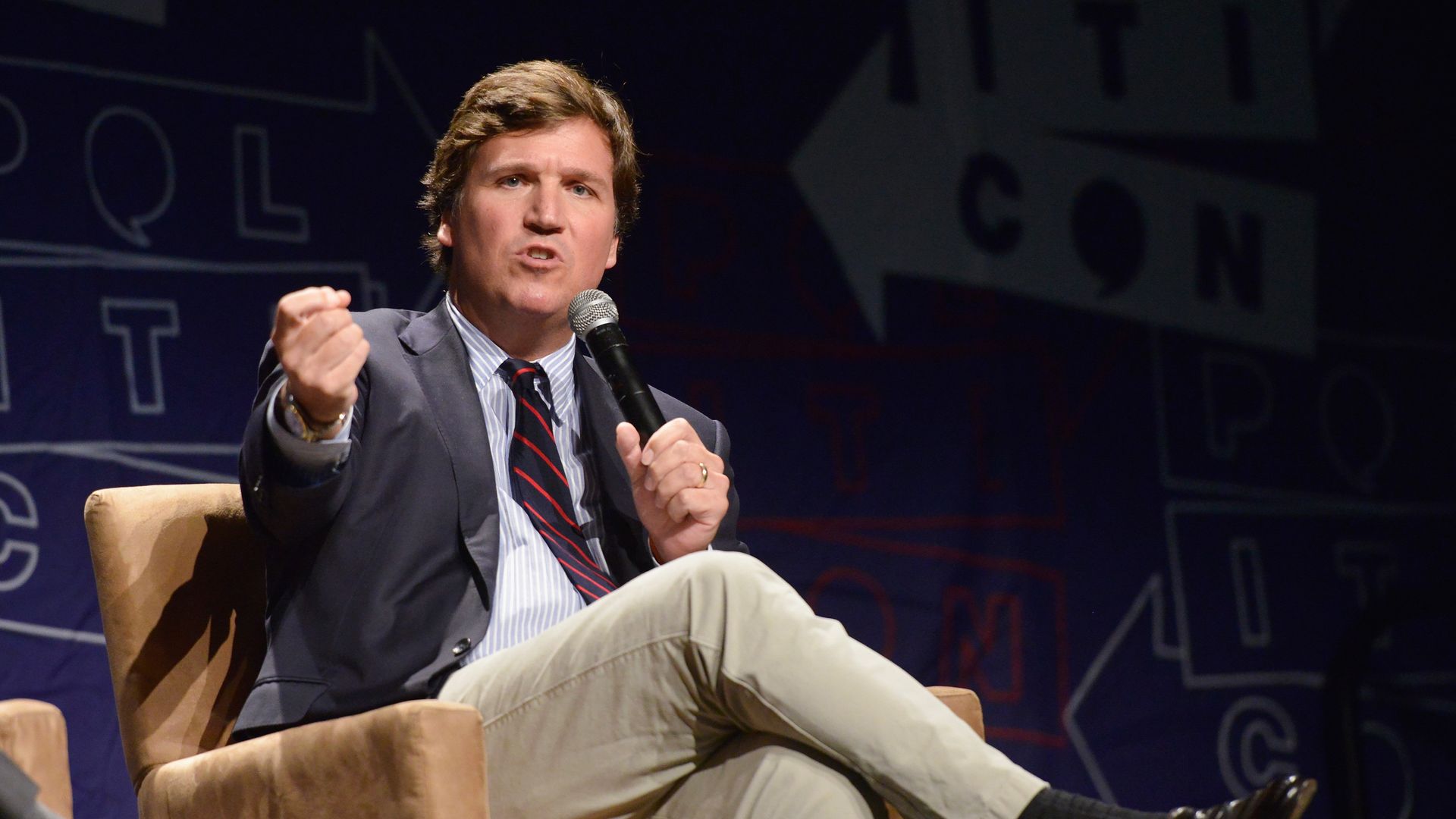 Photo of Tucker Carlson speaking into a microphone with one fist raised as he sits cross-legged in a one-person couch on a stage