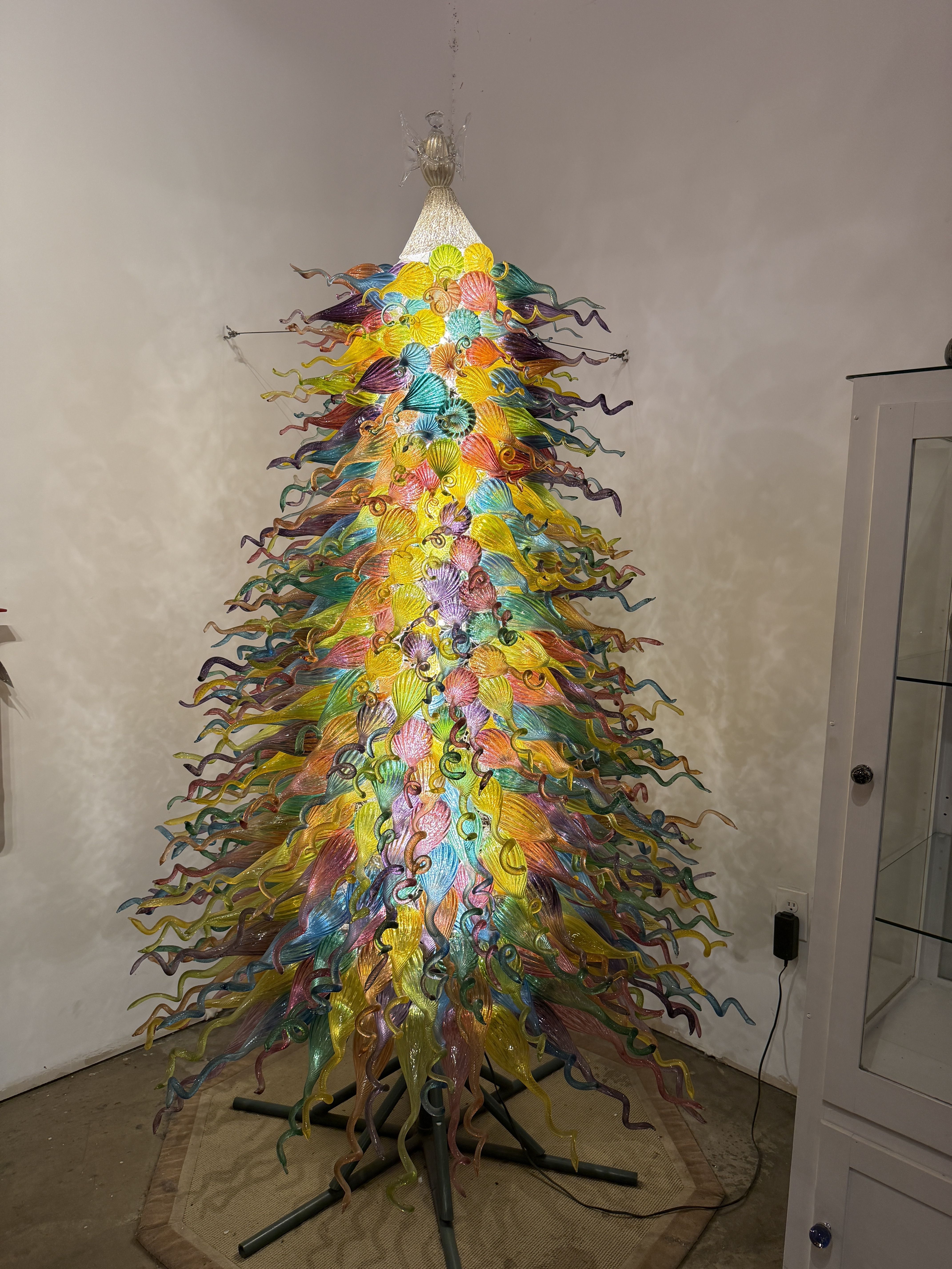 Vibrant glass sculpture shaped like a tree, with rainbow-colored twisting ribbons forming a cone around a central core; mounted on a metal stand in a room with beige walls.