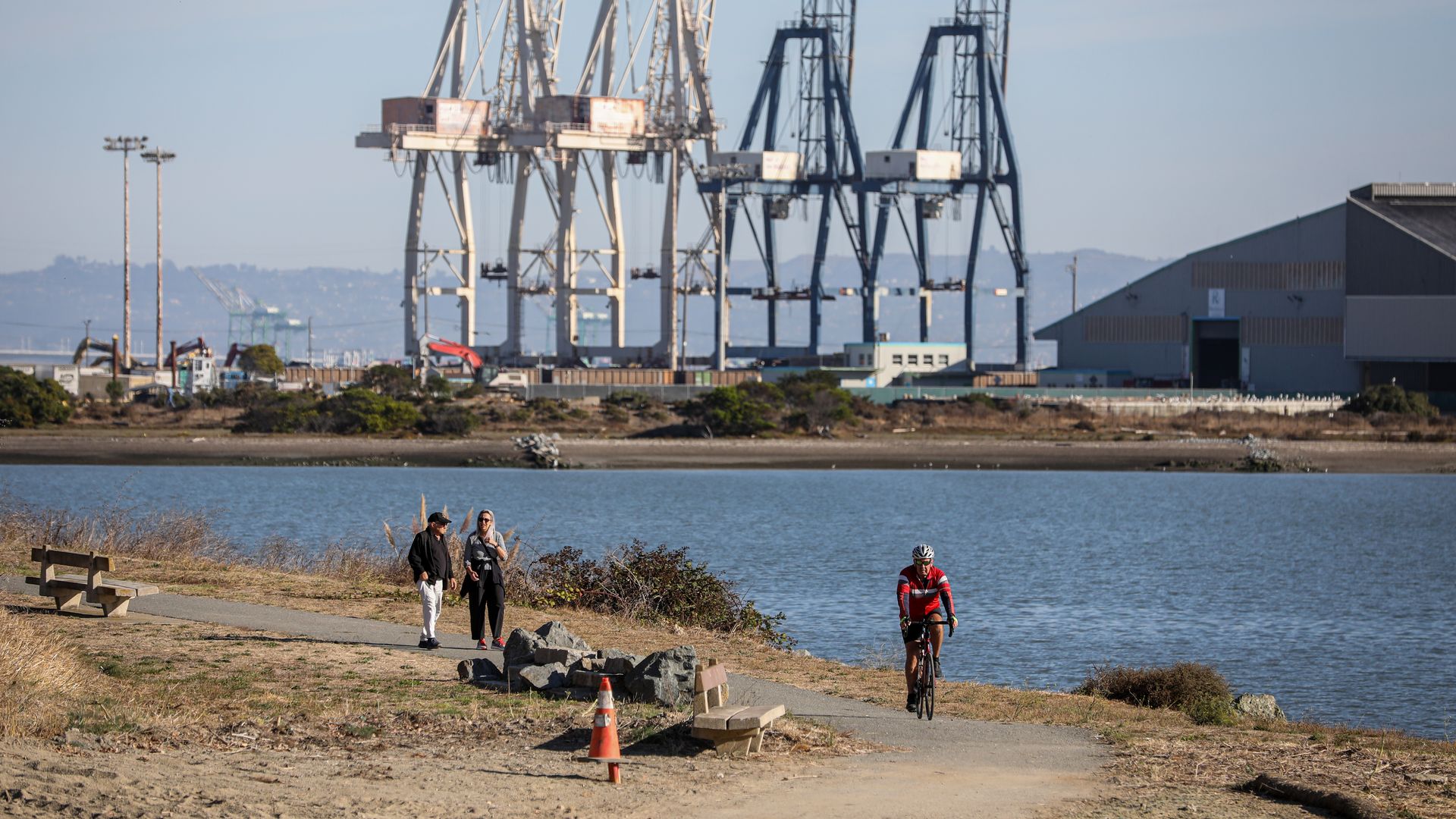 Two people walking along a riverside path with a cyclist in a red jacket riding towards them. Large industrial cranes and a warehouse are visible across the water under a blue sky.