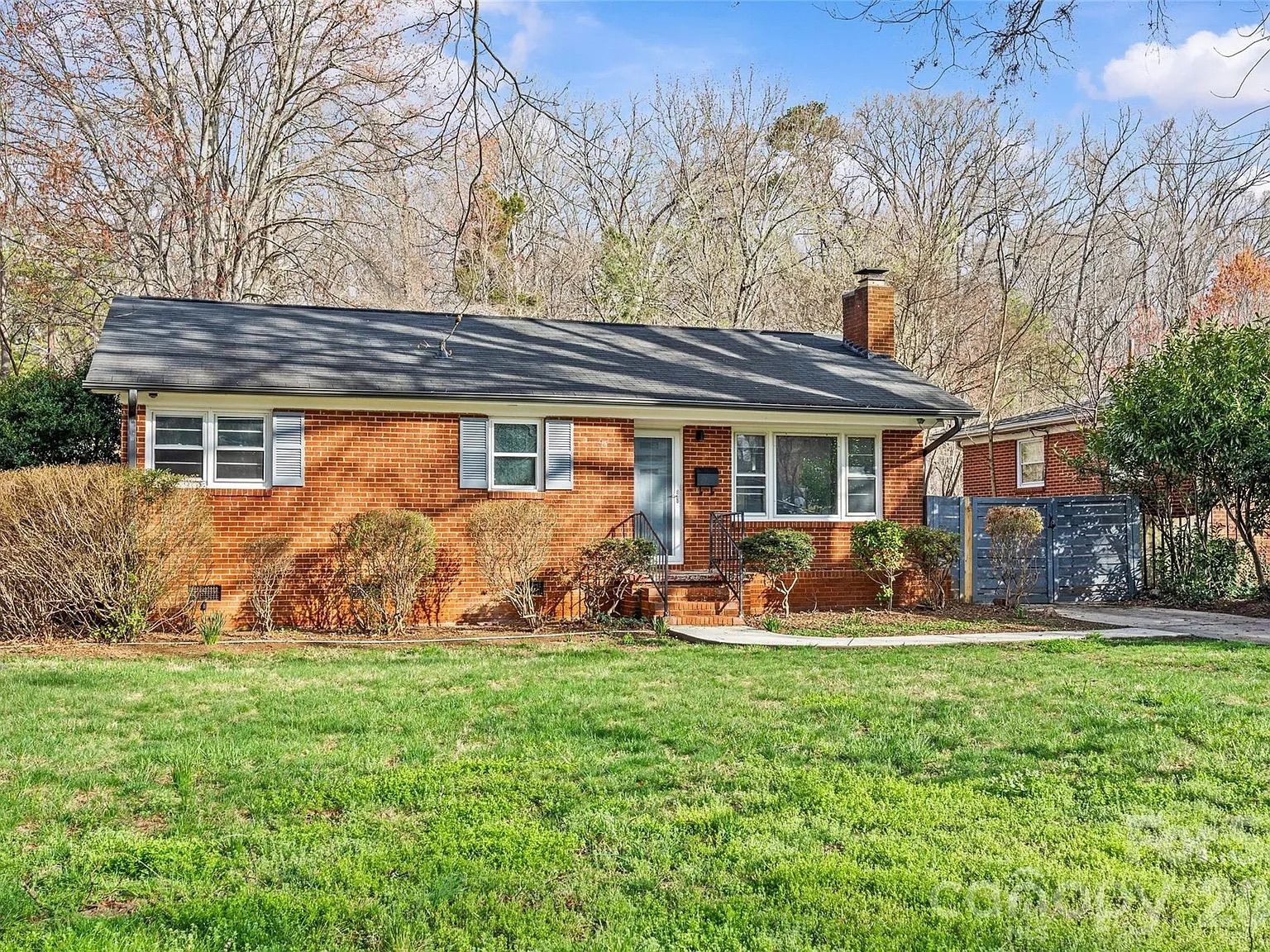 Single-story red brick house with a gray roof and white shutters. Front steps lead to a door; trimmed shrubs line the front yard. Green lawn, bare trees in the background, with a blue gate on the right.