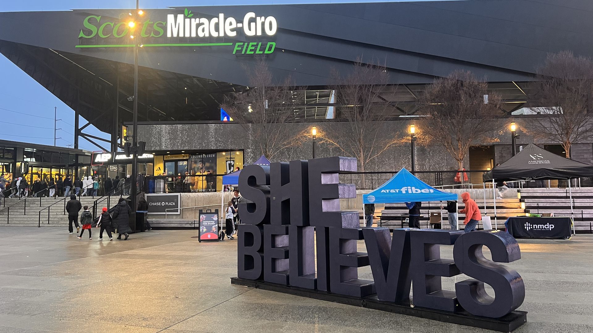 Outdoor plaza at Scotts Miracle-Gro Field in evening light with large black 3D letters spelling "SHE BELIEVES" and people walking near tents and building entrance.
