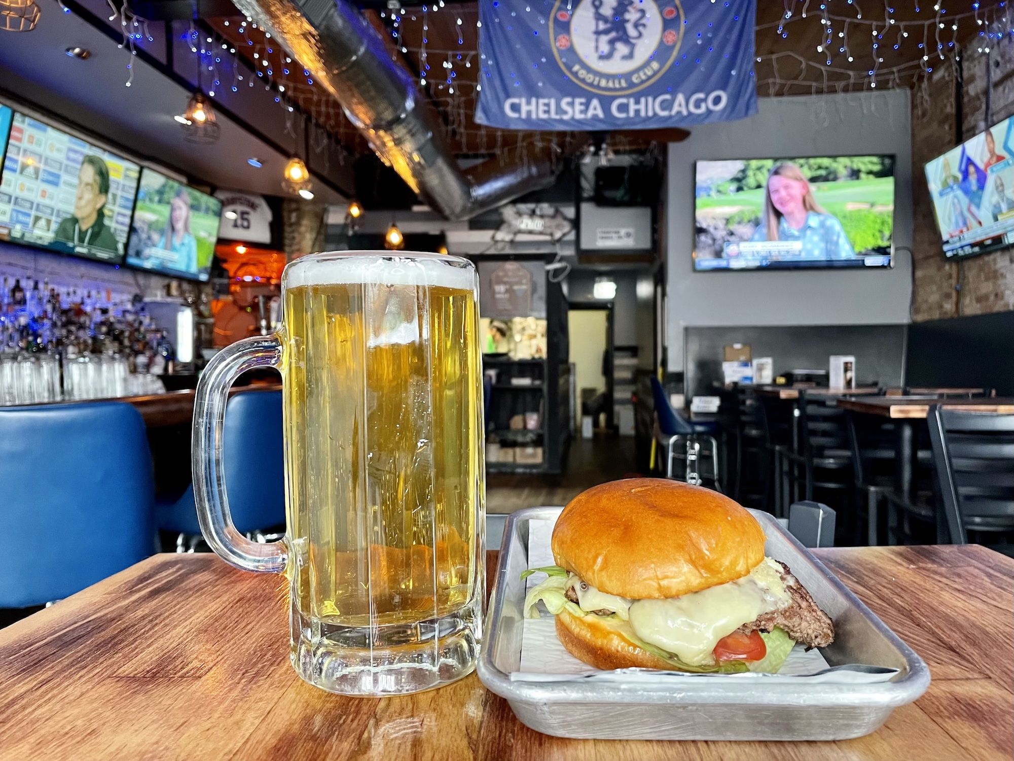 Beer mug and cheeseburger on a wooden table inside a busy sports bar, with multiple TV screens, string lights, and a Chelsea Chicago banner overhead.
