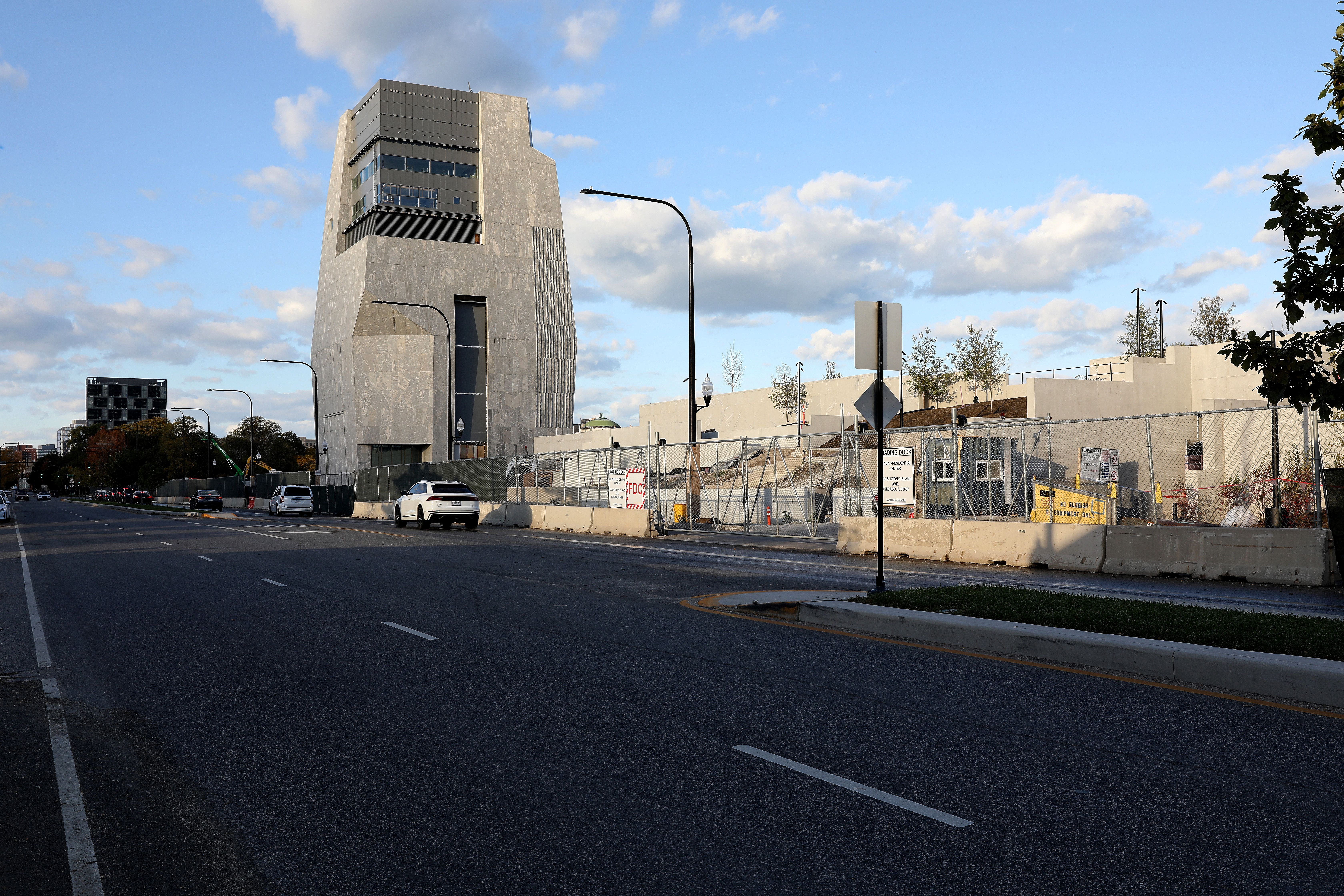 Urban street with a unique, angular gray building under a partly cloudy blue sky. Chain-link fence and construction site with concrete barriers and signs beside the building.
