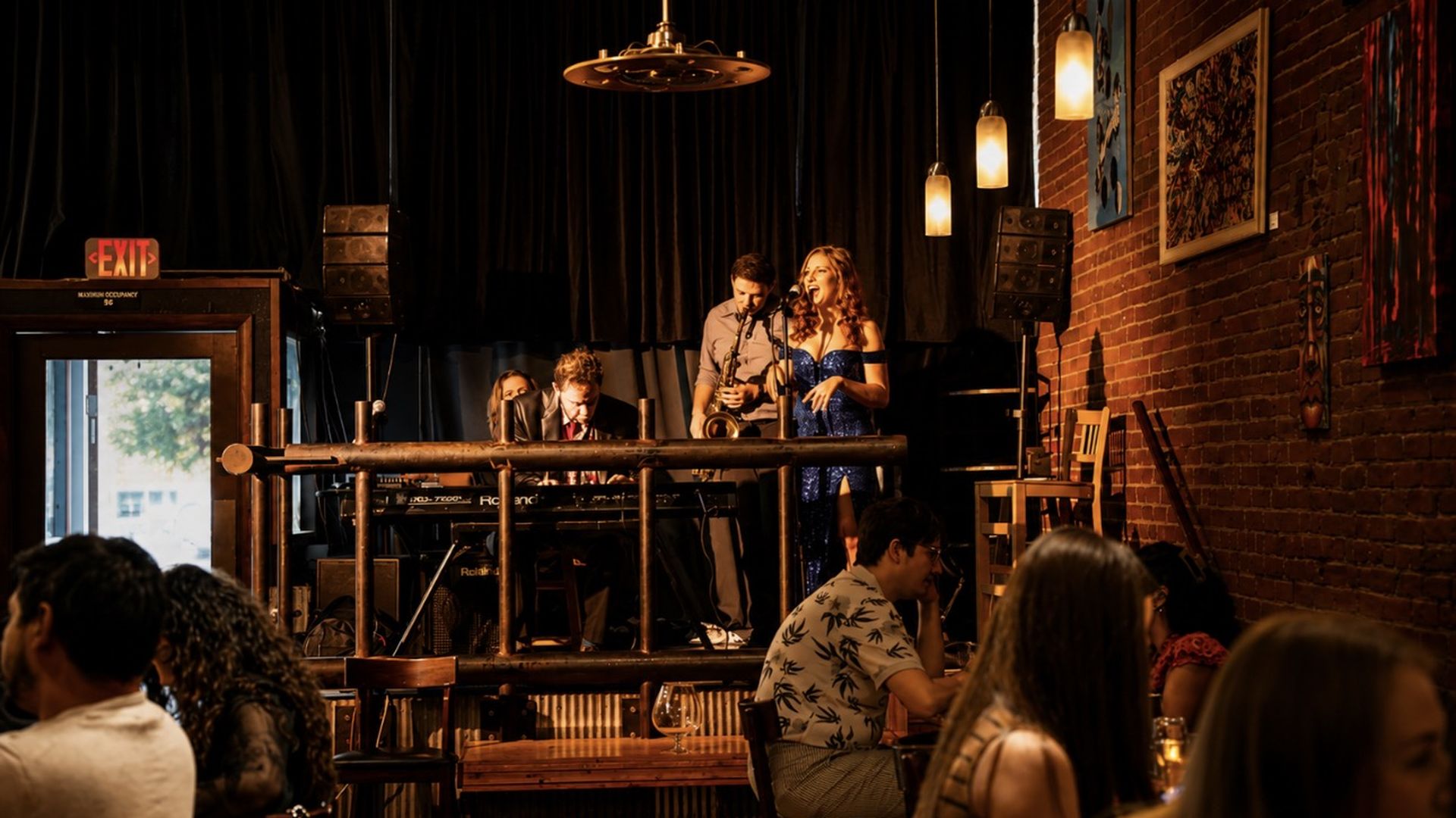 Live band on a brick-walled bar stage: a woman in a blue sparkly dress sings into a mic while two male musicians accompany; warm pendant lights and seated audience in the foreground.
