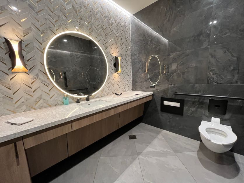 A restroom with lighted round mirror and long vanite with one sing. A white toilet and gray tiled backsplash.