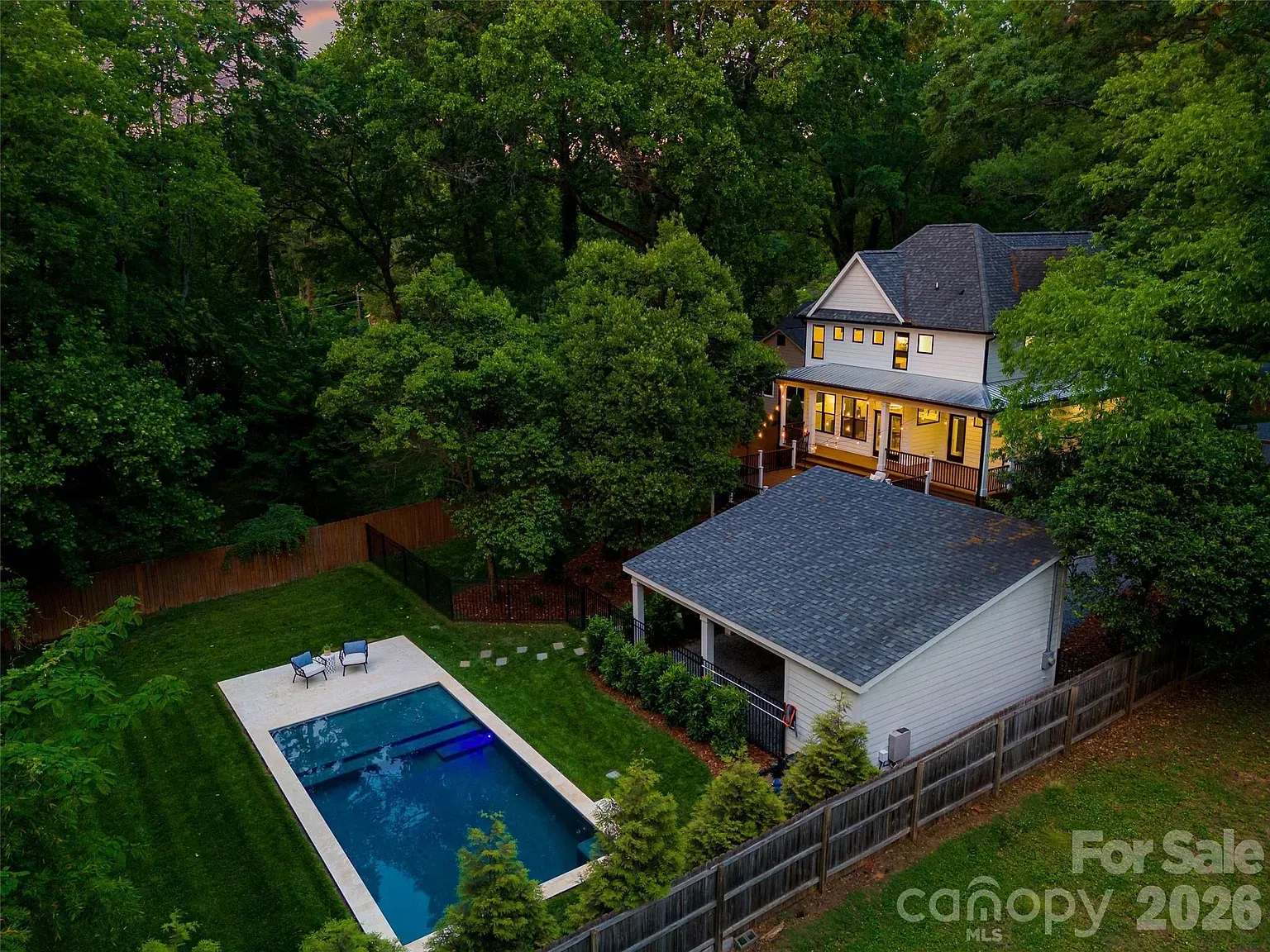 A two-story white house with a wraparound porch sits among dense green trees. A rectangular in-ground pool with blue water sits on a concrete deck in the backyard, fenced off by a wooden fence.