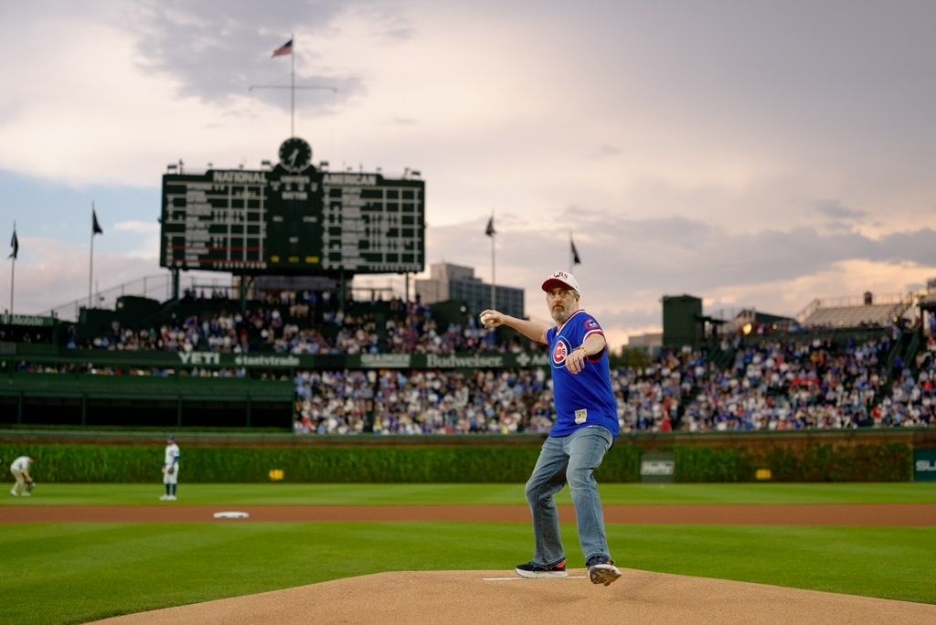 A man in a blue Cubs jersey on the pitcher's mound winds up to pitch, as a full baseball stadium and large scoreboard loom in the background at sunset.