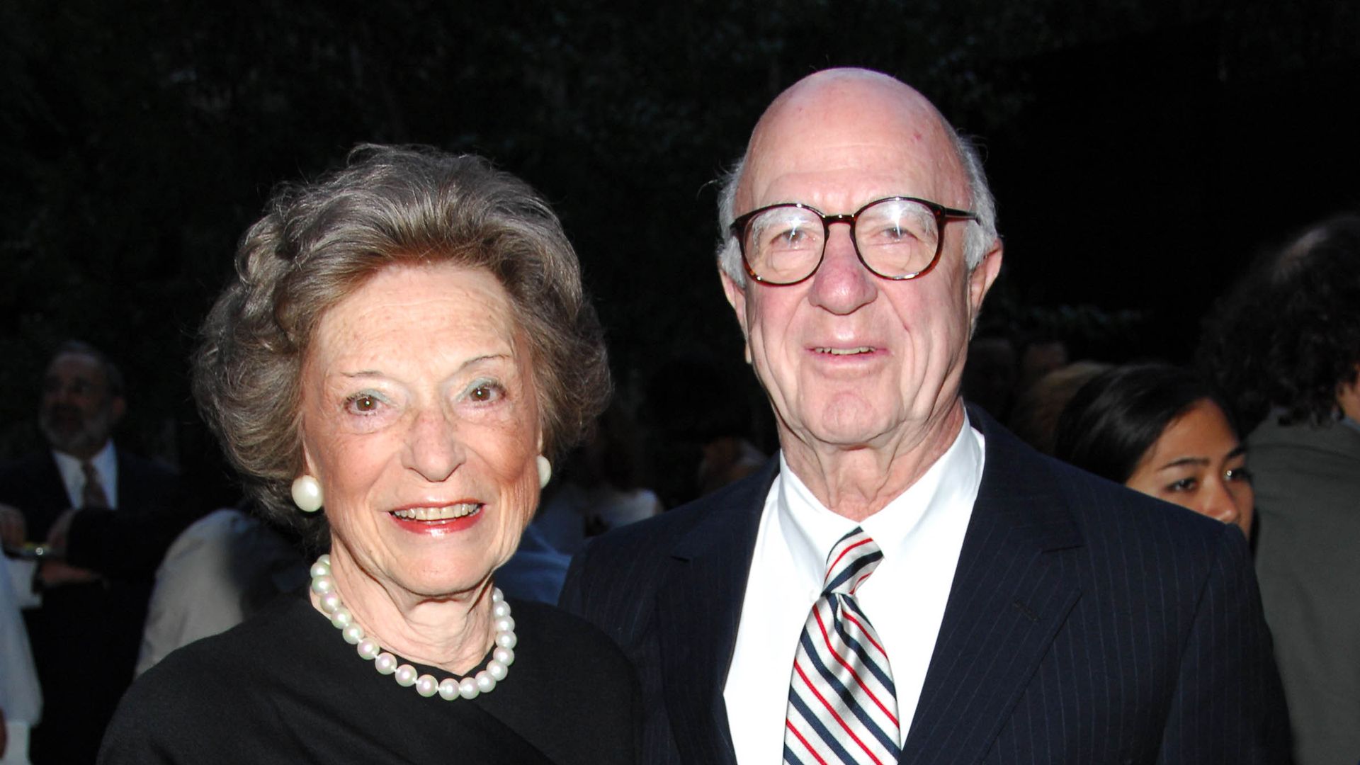 An elderly couple posing at a formal event: a smiling woman in a black dress with a pearl necklace and earrings, and a smiling man in a dark pinstripe suit with a white shirt and striped tie.