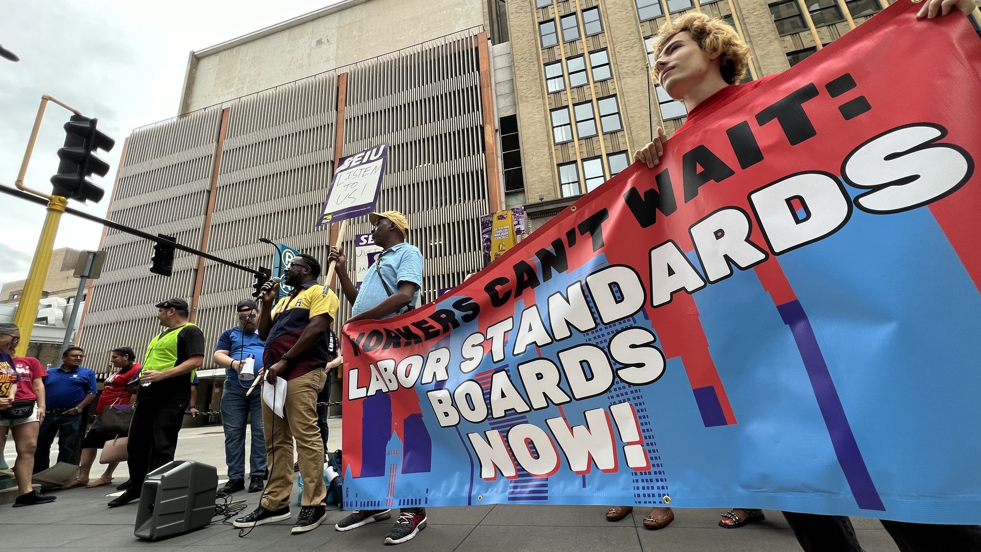 People gathered at a press conference hold a large multi-colored banner reading "Workers Can't Wait: Labor Standards Boards Now!"