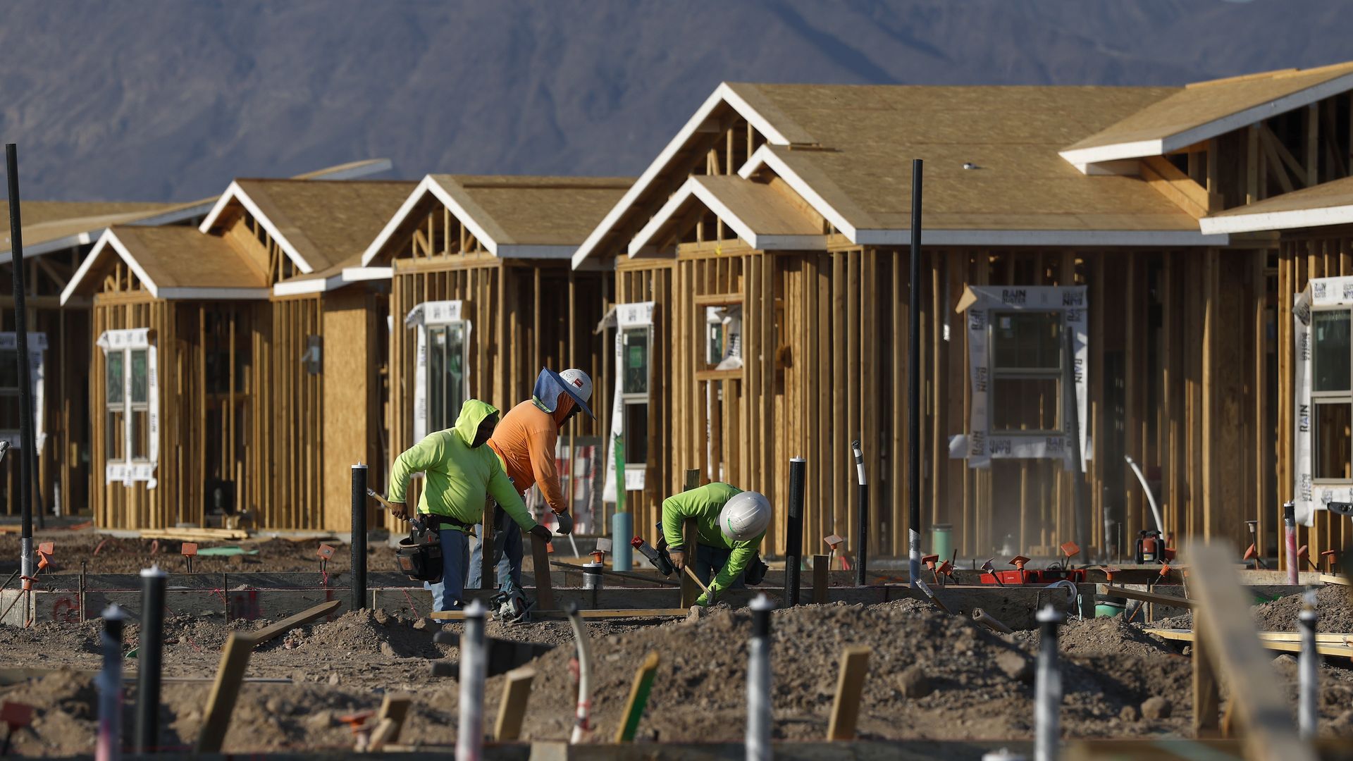 Three construction workers in bright green and orange shirts building the foundation of new wooden houses at a sunny construction site with mountains in the background.