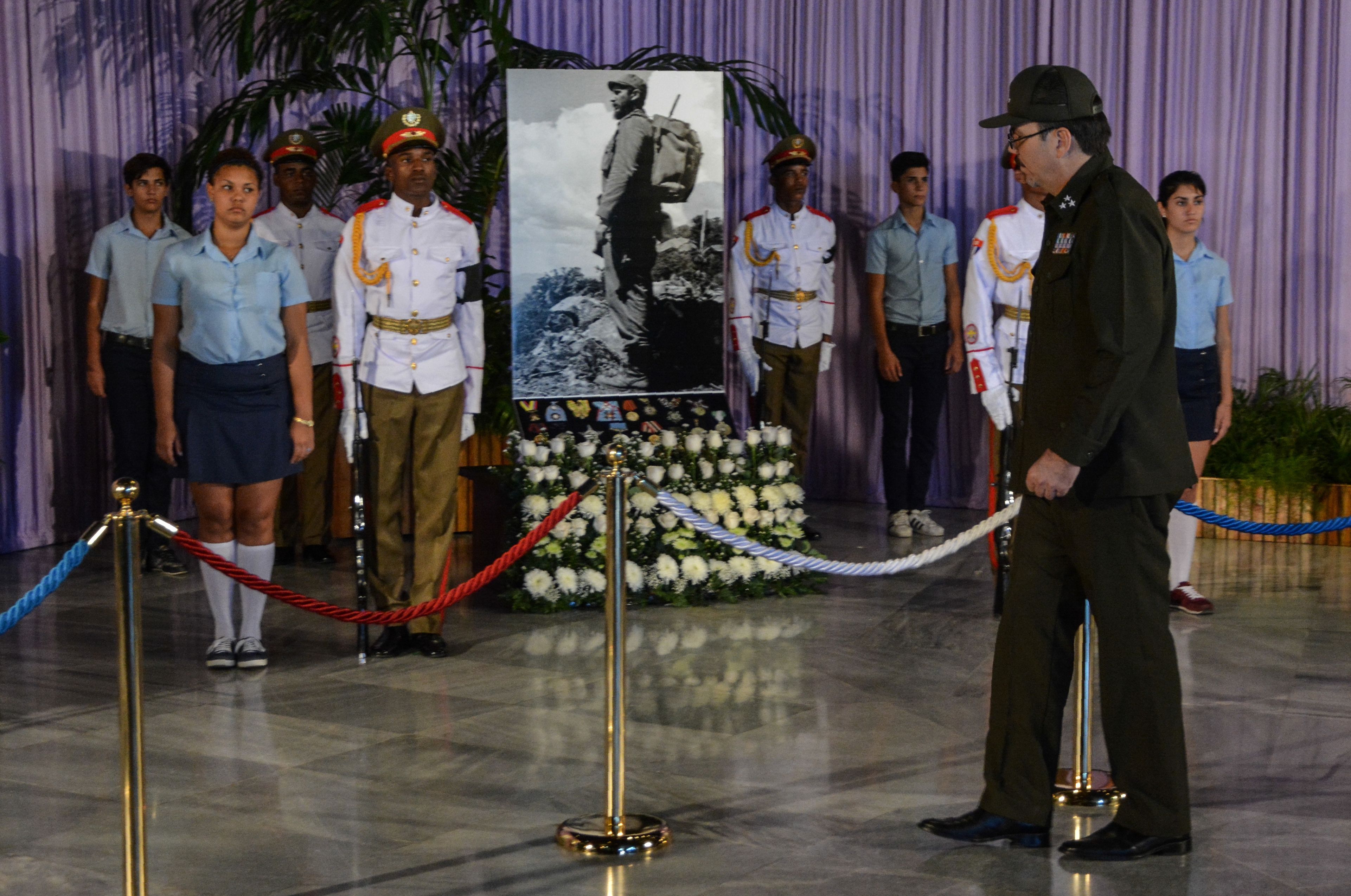 Memorial ceremony with uniformed guards and students standing before a large portrait of a soldier with backpack; white flowers, red and blue ropes, and a purple backdrop.