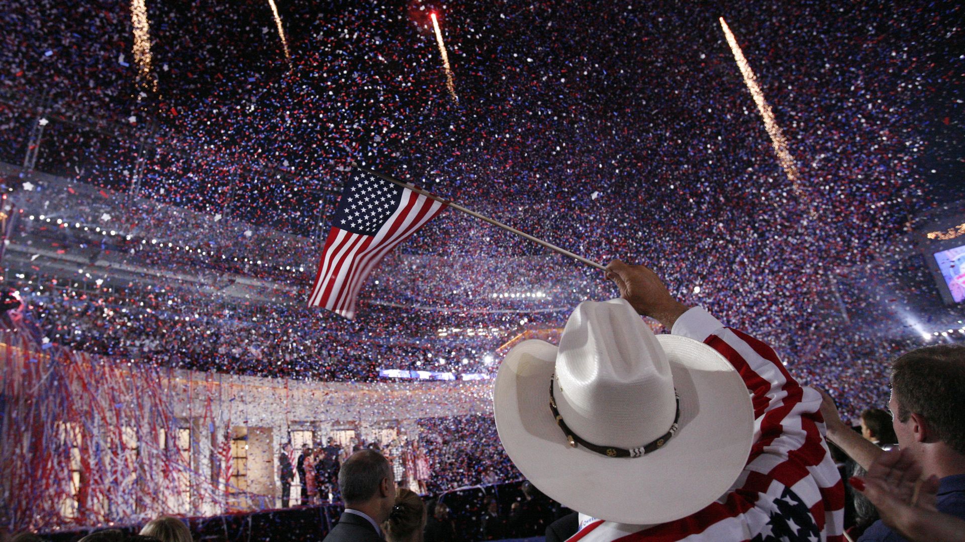 Crowded stadium at night as fireworks and confetti rain down; a person in a white cowboy hat waves an American flag amid cheering fans in red, white and blue.