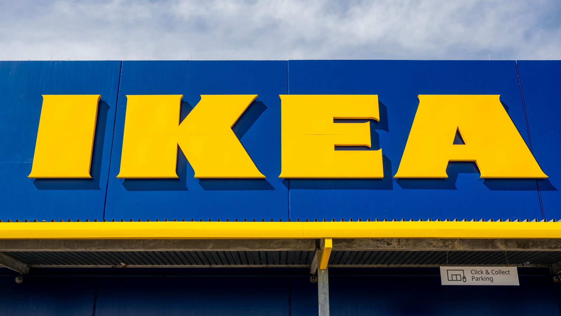 Exterior of an IKEA store with a bold yellow IKEA sign on a blue wall, clear sky above, and a small Click & Collect Parking sign beneath.
