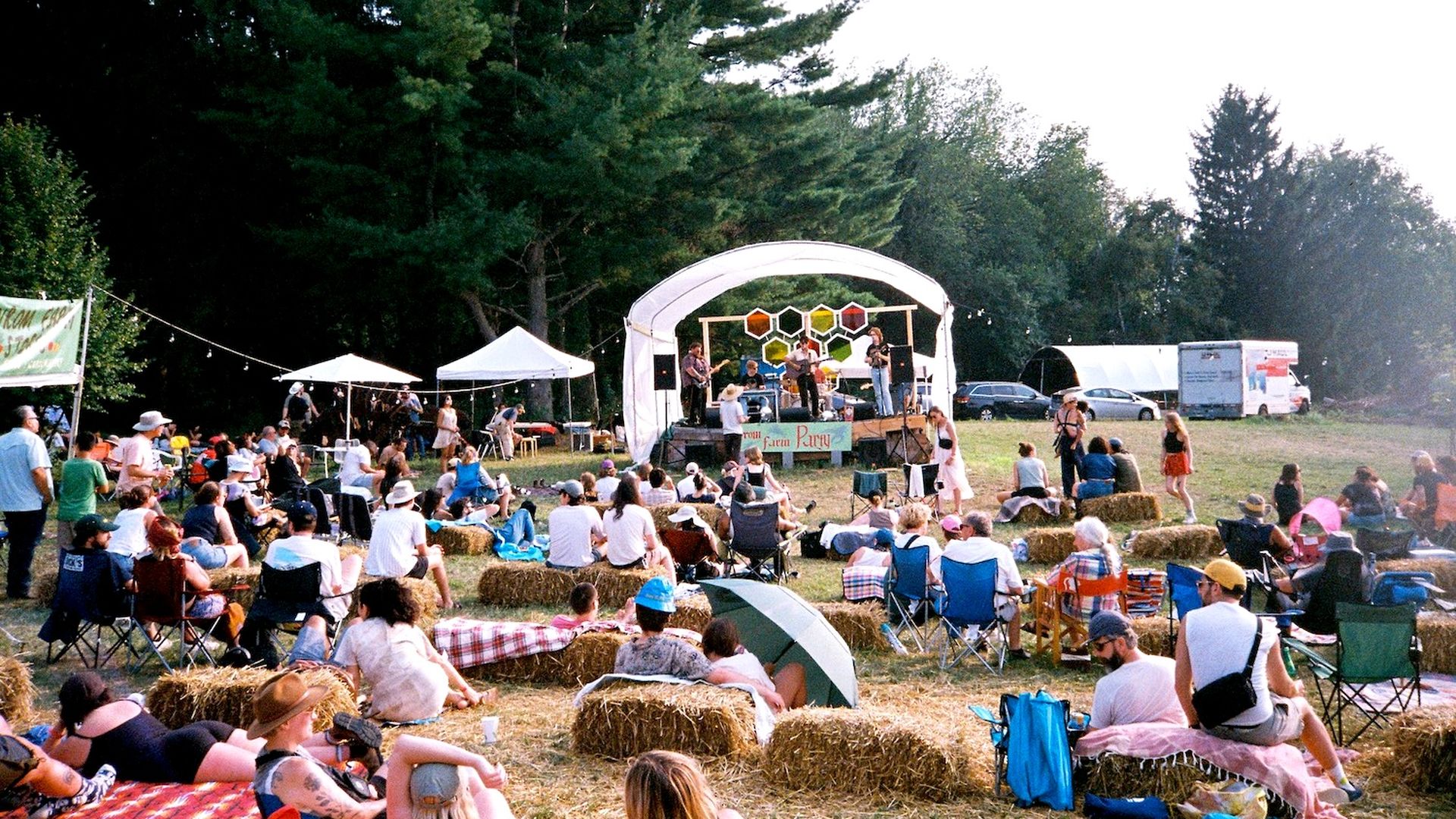 Outdoor music festival in a grassy park: a white arched stage with a honeycomb backdrop, musicians playing; audience sits on hay bales and blankets, with tents and trees surrounding the crowd.