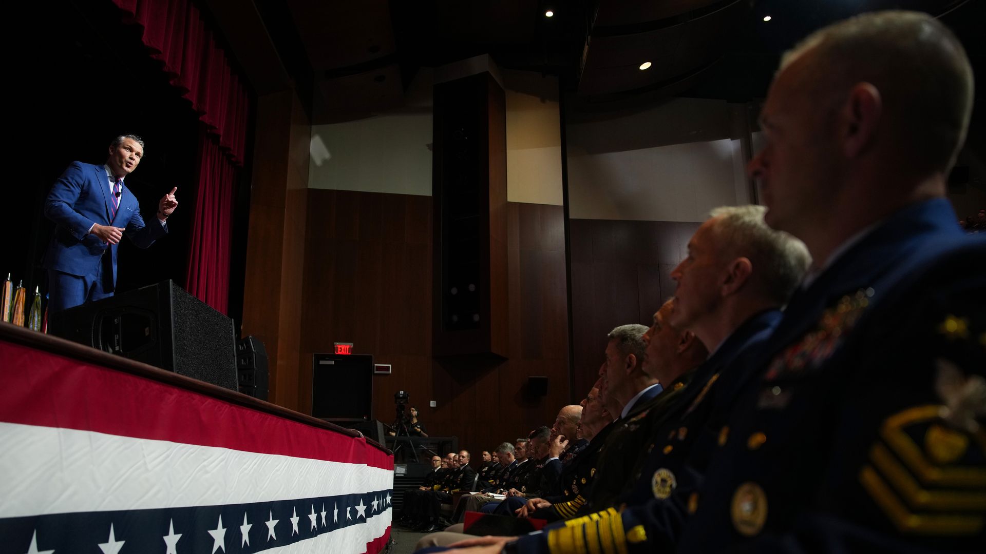 Pete Hegseth, a man in a blue suit, stands on stage and speaks to a crowd of uniformed military members, including admirals and generals. There stars-and-stripes decorations throughout.