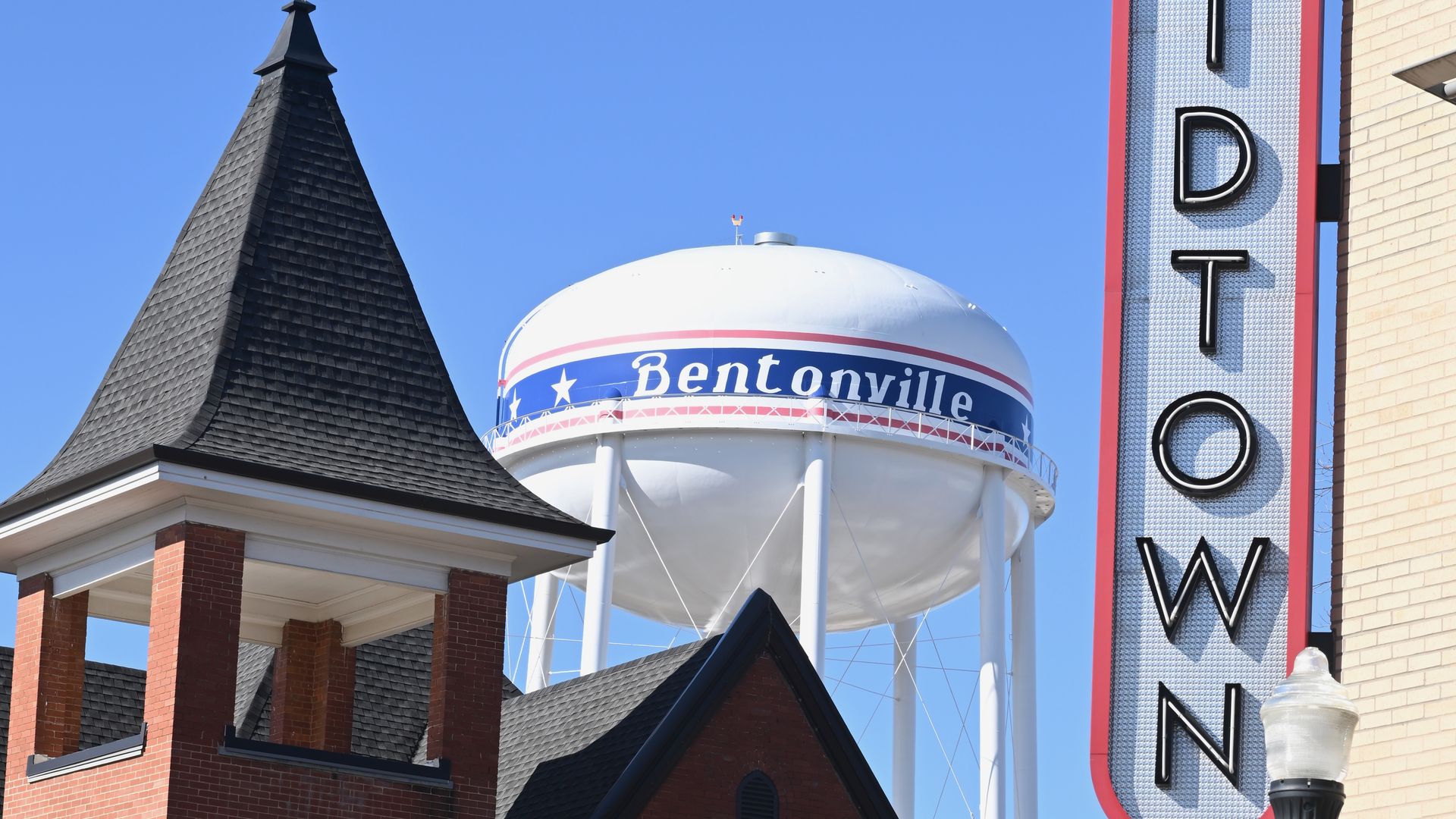 White Bentonville water tower with red and blue stripe and stars behind brick buildings with black roofs and a vertical "DOWNTOWN" sign on a clear blue sky day.
