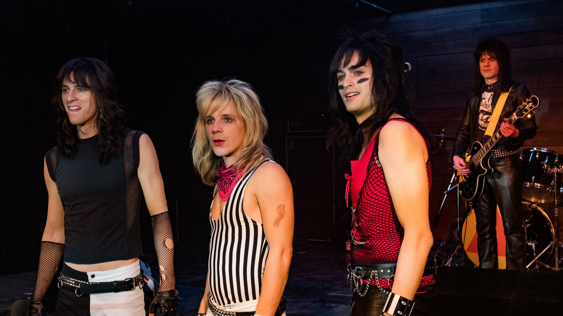 Four rock performers on stage: left in a black sleeveless top; next a blonde in stripes; next a black-haired person with face paint in a red mesh top; far right guitarist in leather.