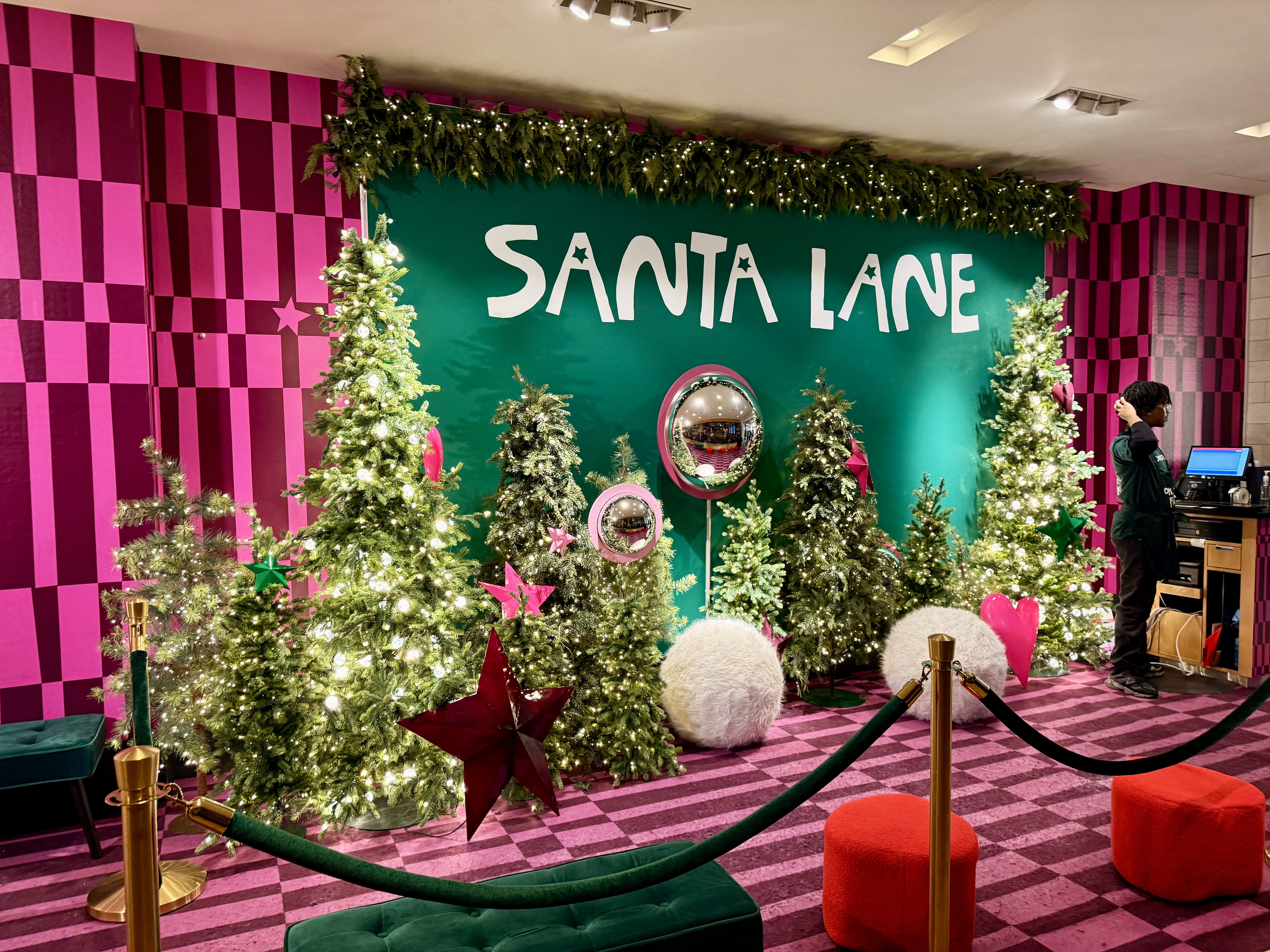 Holiday display with lit Christmas trees decorated with red and green stars, white fluffy ornaments, and heart shapes in front of a green wall with "SANTA LANE" text on a pink checkered background.