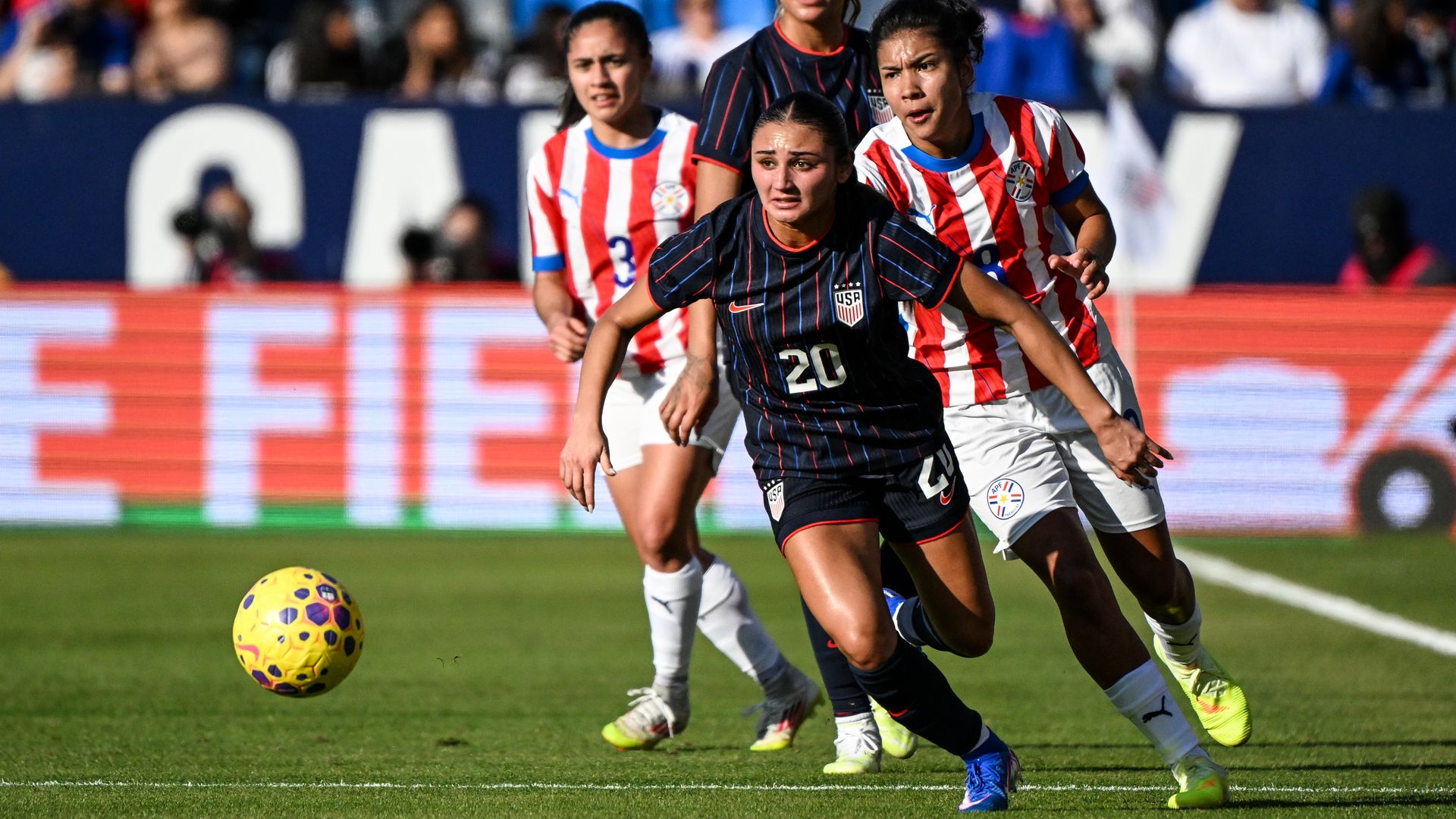 Soccer match scene with four female players sprinting after a yellow ball on a green field; two players wear red-and-white stripes, two wear dark blue jerseys with USA crest; blurred crowd in stands.