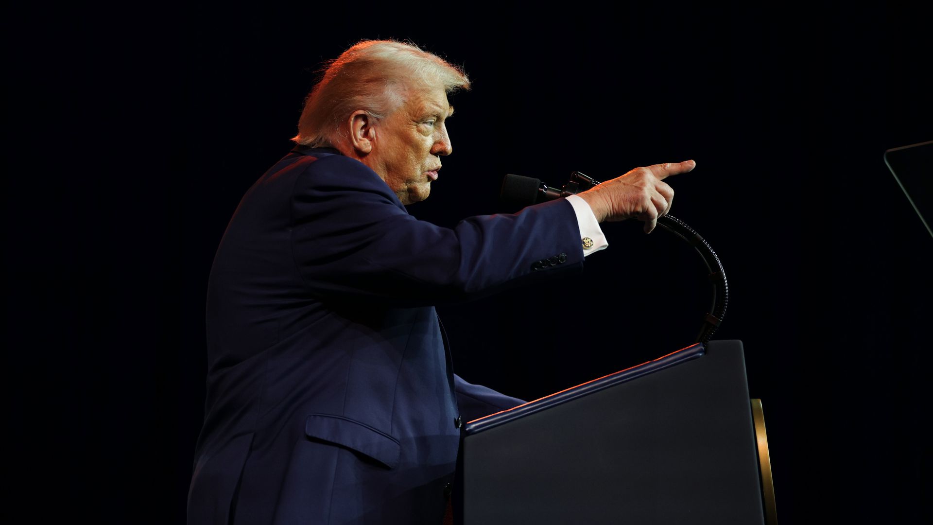 President Trump — wearing a navy suit and a white shirt — points as he speaks into a microphone attached to a podium.