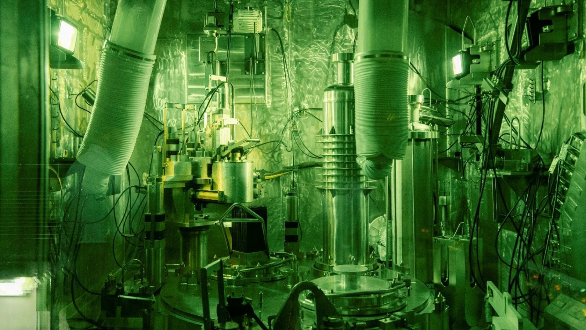Green-lit industrial or scientific lab with metal equipment, tubes, wires, and control panels in a reflective metal-walled room.