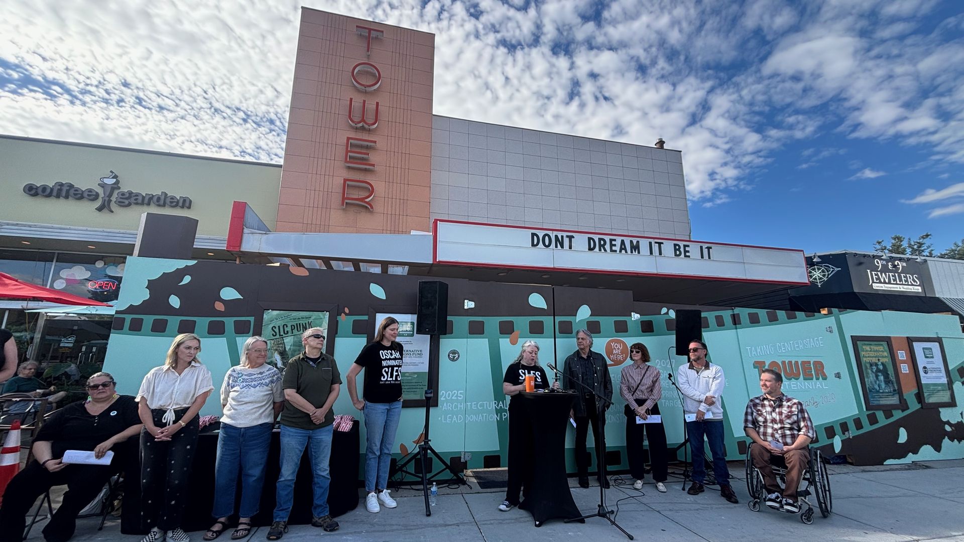 Group of people on a stage outside a building with a tall pink "TOWER" sign and the marquee "DONT DREAM IT BE IT." Left shows a "coffee garden" sign; blue sky with scattered clouds.