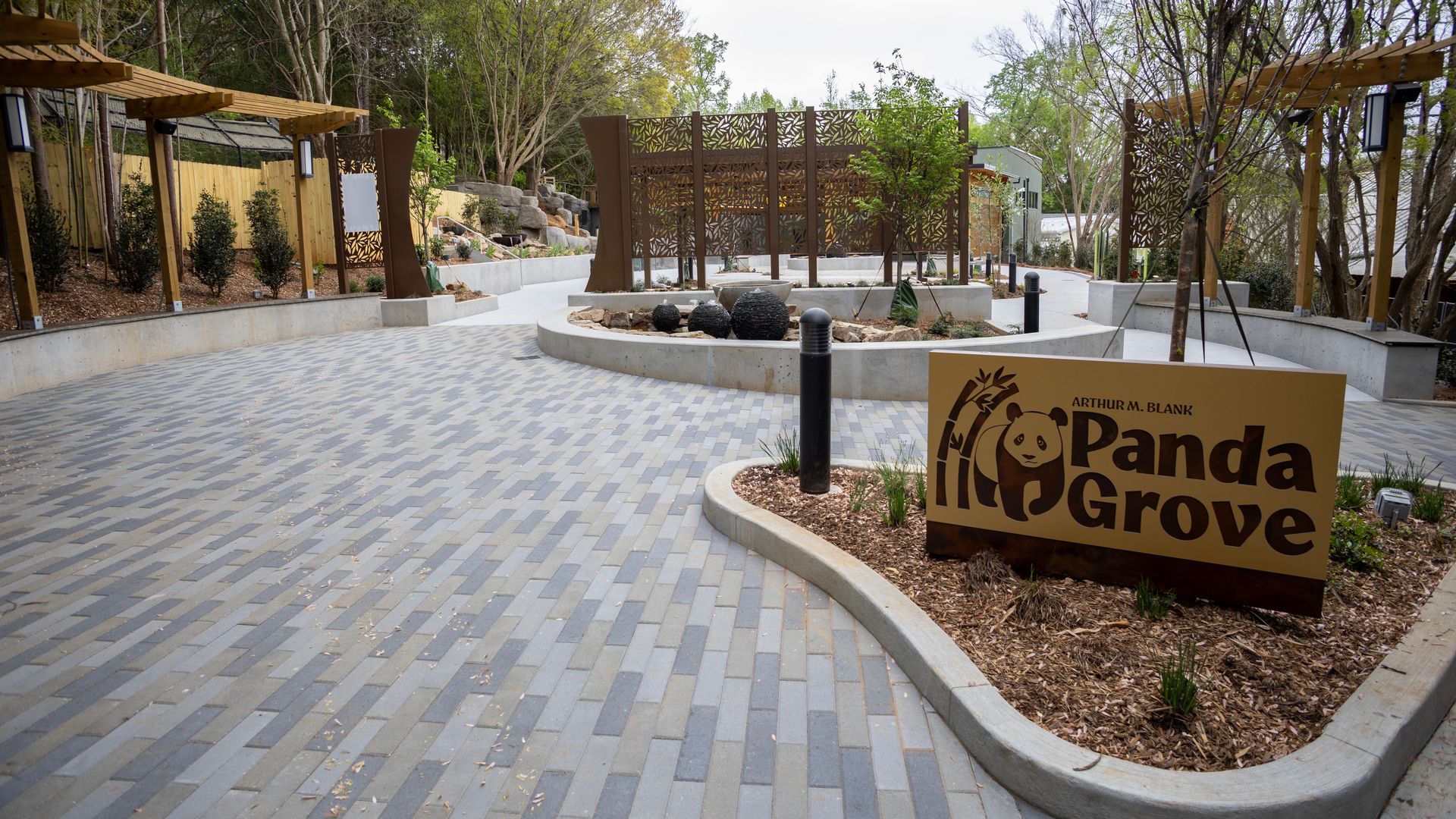 Wide courtyard with gray-blue brick paving, circular planters, and brown screens. Wooden pergolas line the space; landscaping with rocks and a central fountain of black spheres, plus a yellow sign reading Panda Grove.