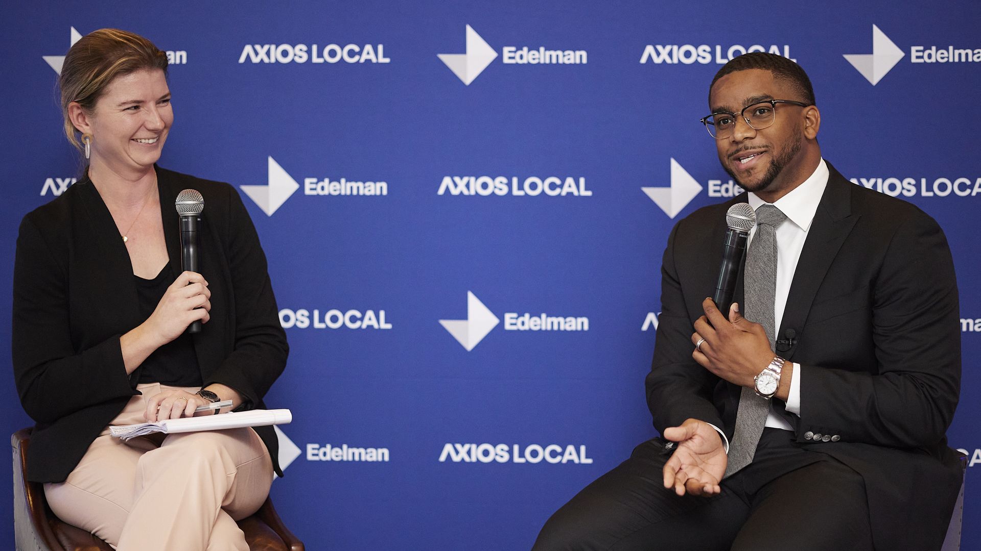 A woman holds a microphone and laughs while a man wearing a suit speaks and smiles. Behind them a screen reads Axios Local and Edelman