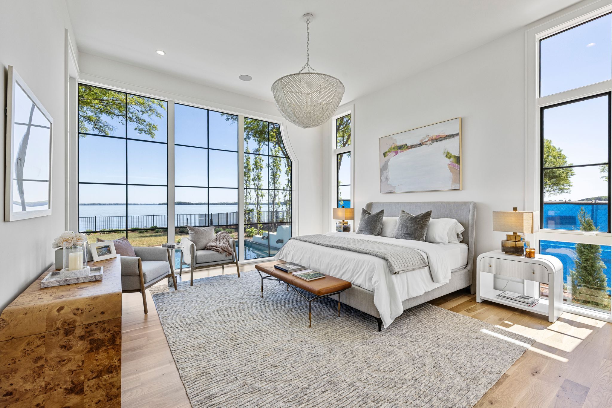 Modern bedroom with large windows overlooking a lake, featuring a gray bed with white bedding, two gray armchairs, a wooden dresser, a bench, lamps, and light hardwood floors.