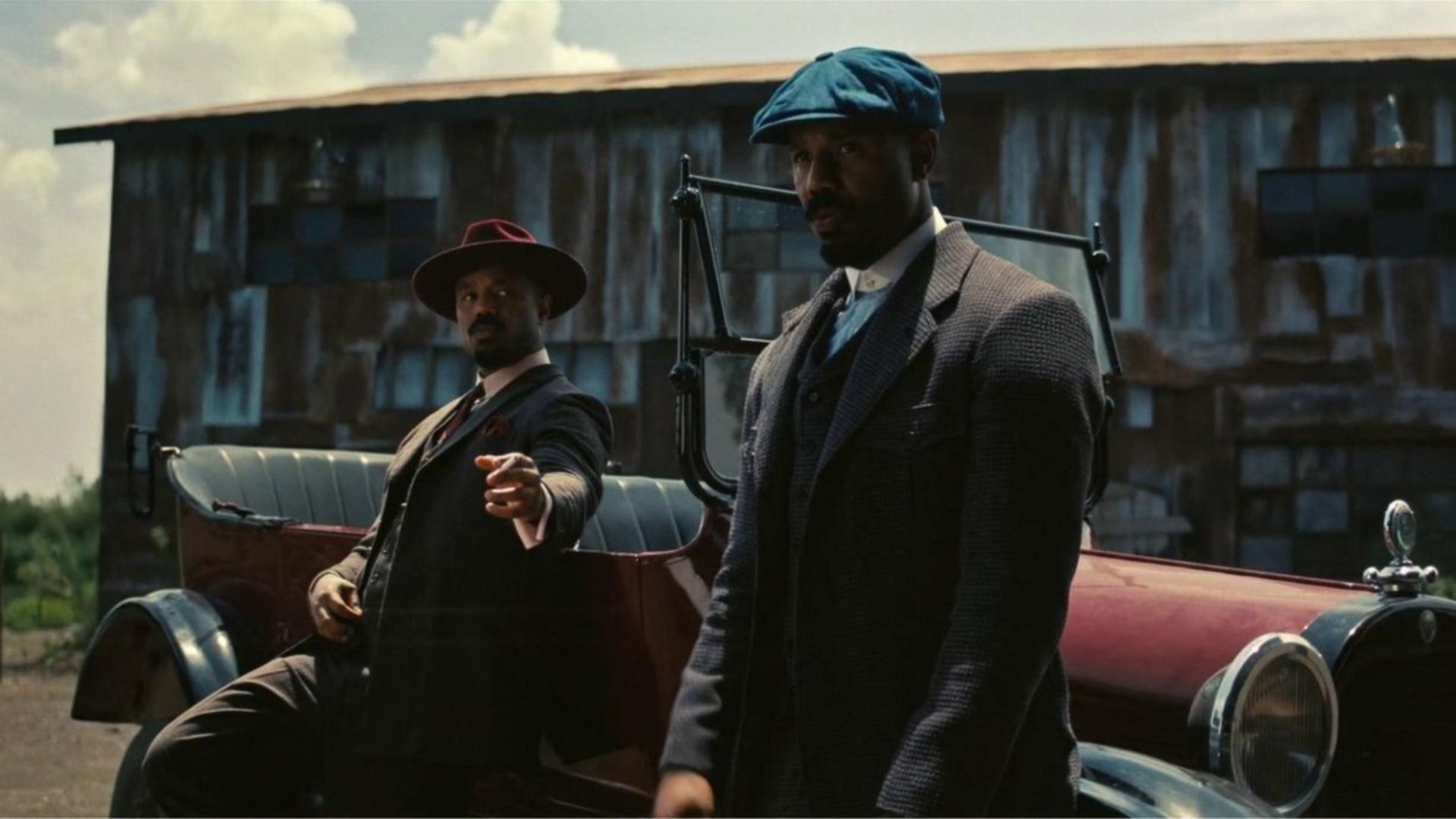 Two men in vintage attire stand by a red classic car next to a weathered wooden building. One wears a blue cap and checkered coat; the other a red hat, pointing toward the camera.