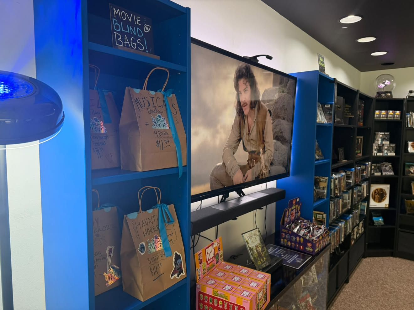Blue bookstore display with a large TV showing a man in a tan outfit in a desert scene; brown paper bags labeled "Movie Blind Bags" rest on shelves beside games and books.