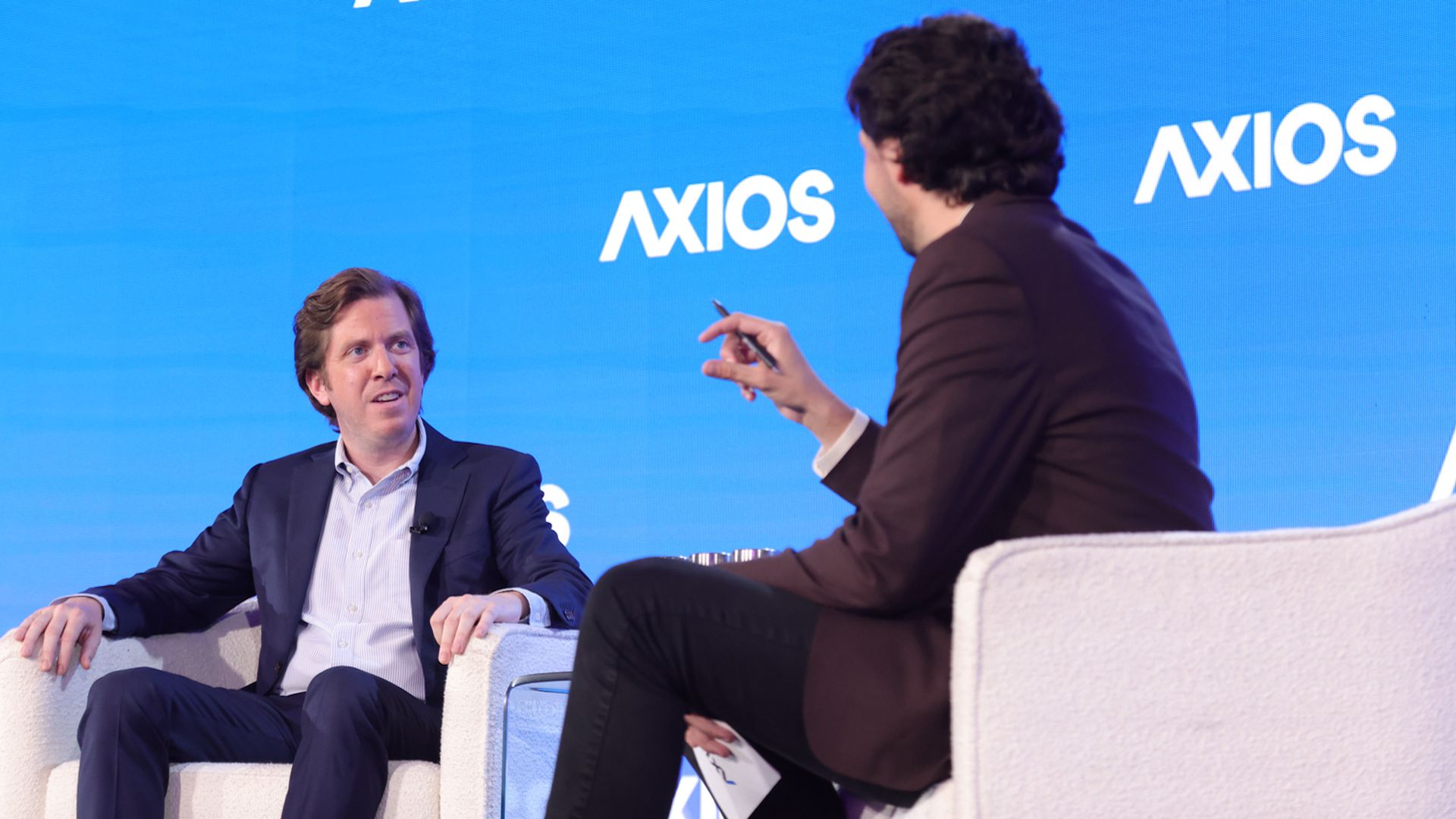 Two men in suits seated on white chairs having a discussion on a stage with a blue background displaying the word "AXIOS" multiple times.
