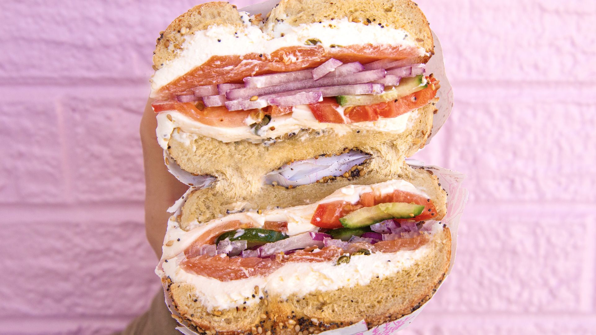 Close-up of a multi-layer bagel sandwich with cream cheese, smoked salmon, red onions, tomatoes and cucumbers, sprinkled with sesame seeds, held against a pink textured background.