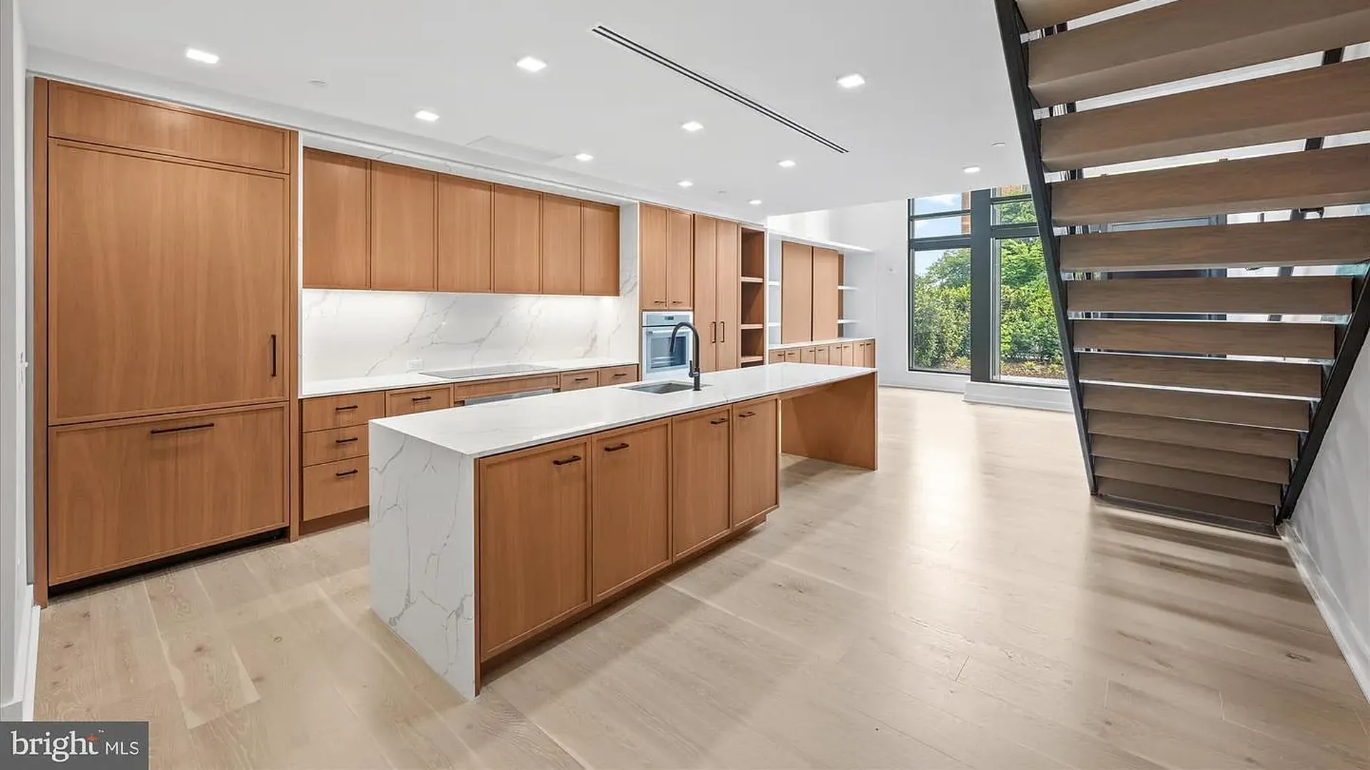 Modern kitchen with warm wood cabinetry, white marble countertops and an island sink. Built-in oven, wide window, light wood floors, and a dark metal-and-wood staircase on the right.