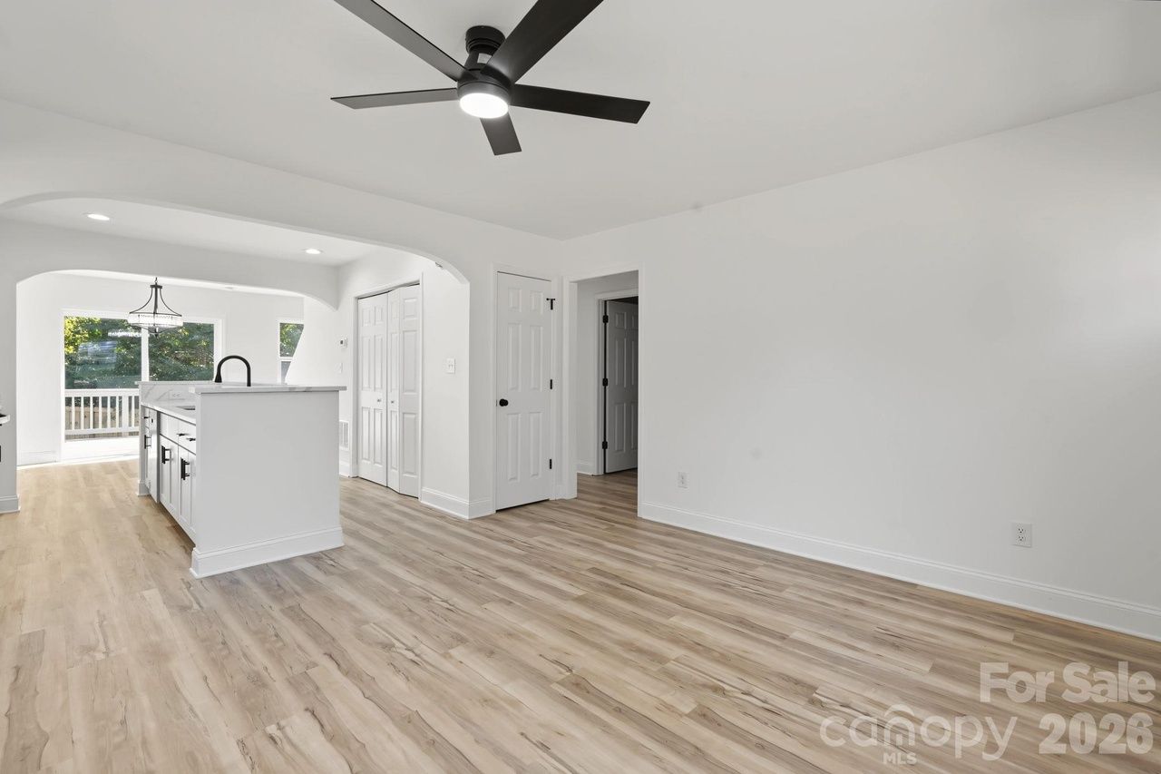 Bright open living area with light wood flooring, white walls, and a black ceiling fan. A white kitchen island with a sink stands near an arched opening to the kitchen and a deck door.