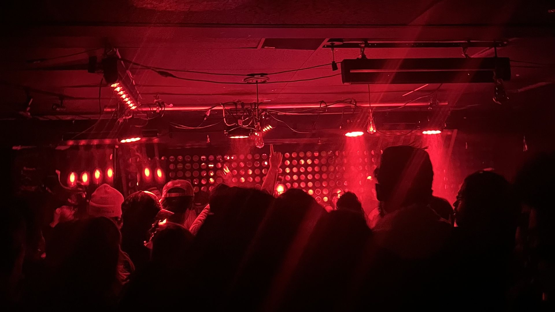 Dark nightclub or concert venue bathed in red light; silhouettes of a crowded audience facing a glowing LED wall, with ceiling fixtures and cables visible above.