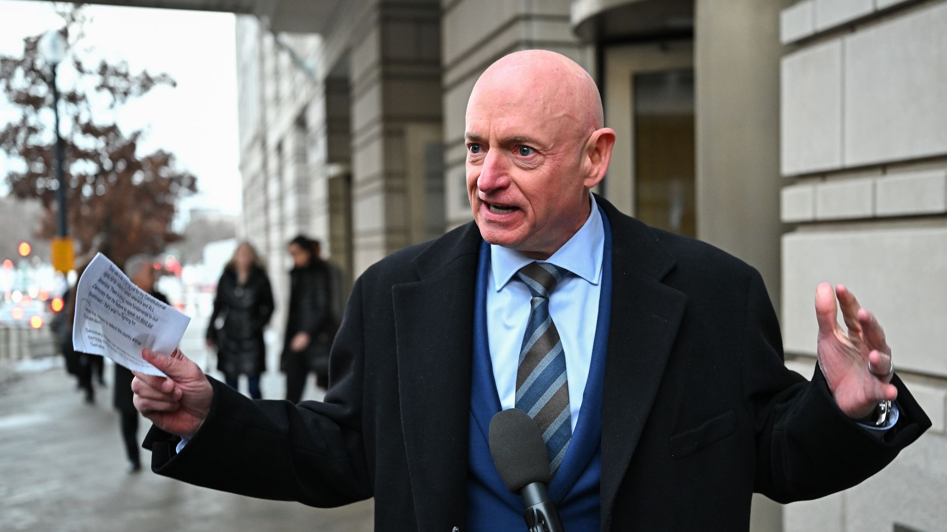Bald man in a dark suit, blue shirt and striped tie, speaks into a microphone while gesturing with his hands on a city sidewalk; he holds a sheet of paper, with a concrete building in the background.
