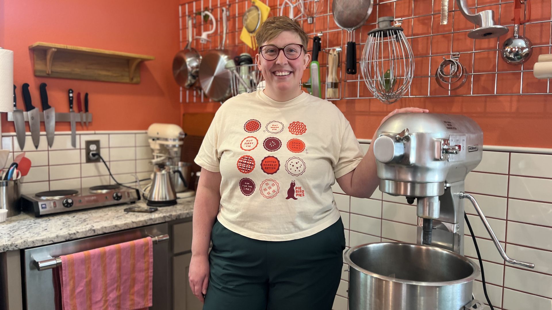 Smiling person with short hair and glasses stands in an orange kitchen, in front of a wall grid with hanging utensils (whisks, ladles, pans). A large mixer sits to their right.