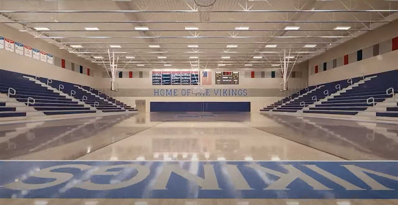 Empty school gym with polished wooden floor, blue bleachers, basketball hoops, scoreboard, and text 'HOME OF THE VIKINGS' on the far wall in blue letters.