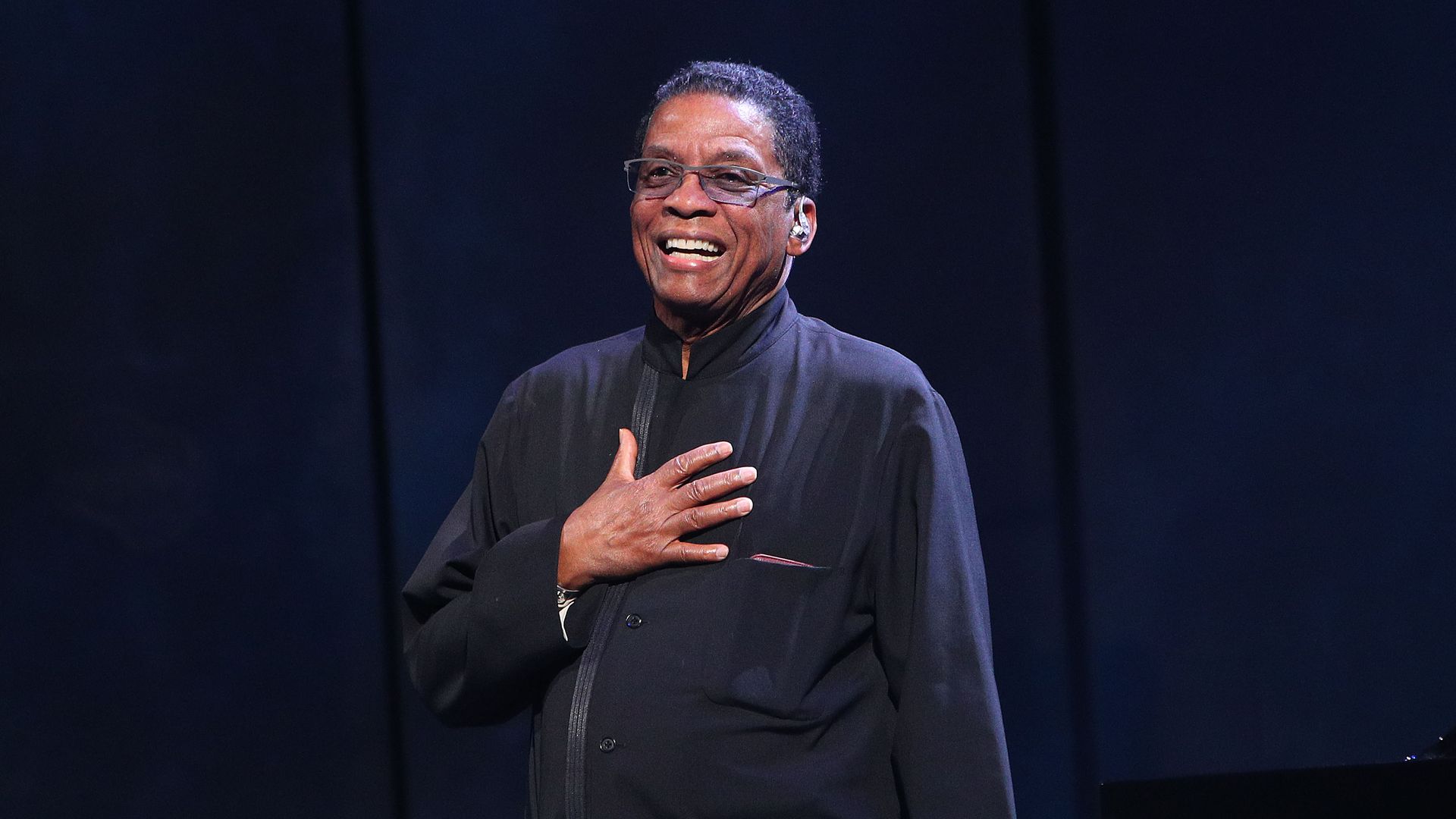 Herbie Hancock stands on stage with his right hand on his chest. 