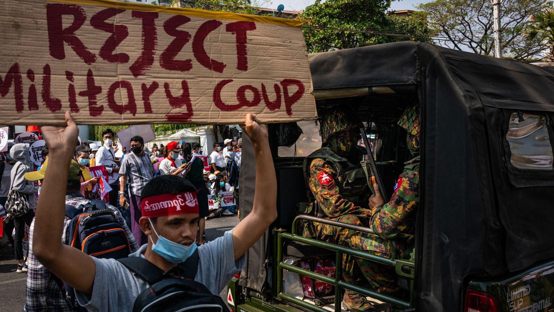 Photo of a protester holding a sign that says "Reject military coup" while walking next to a vehicle of soldiers