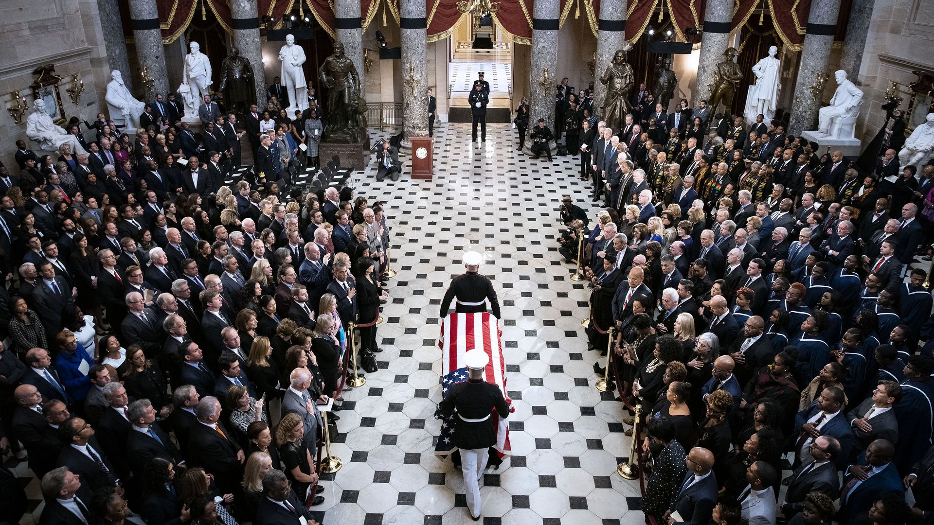 In this image, two soldiers carry a coffin draped with the American flag through a crowd in the Capitol. A path has been cleared for them.