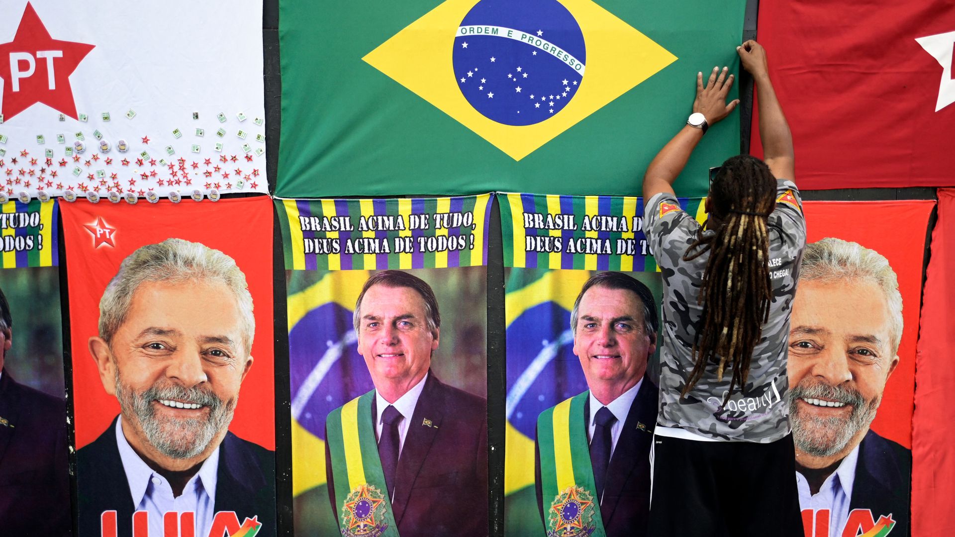 a person pins up a Brazil flag near banners for presidential candidates Jair Bolosonaro Luiz Inácio Lula da Silva