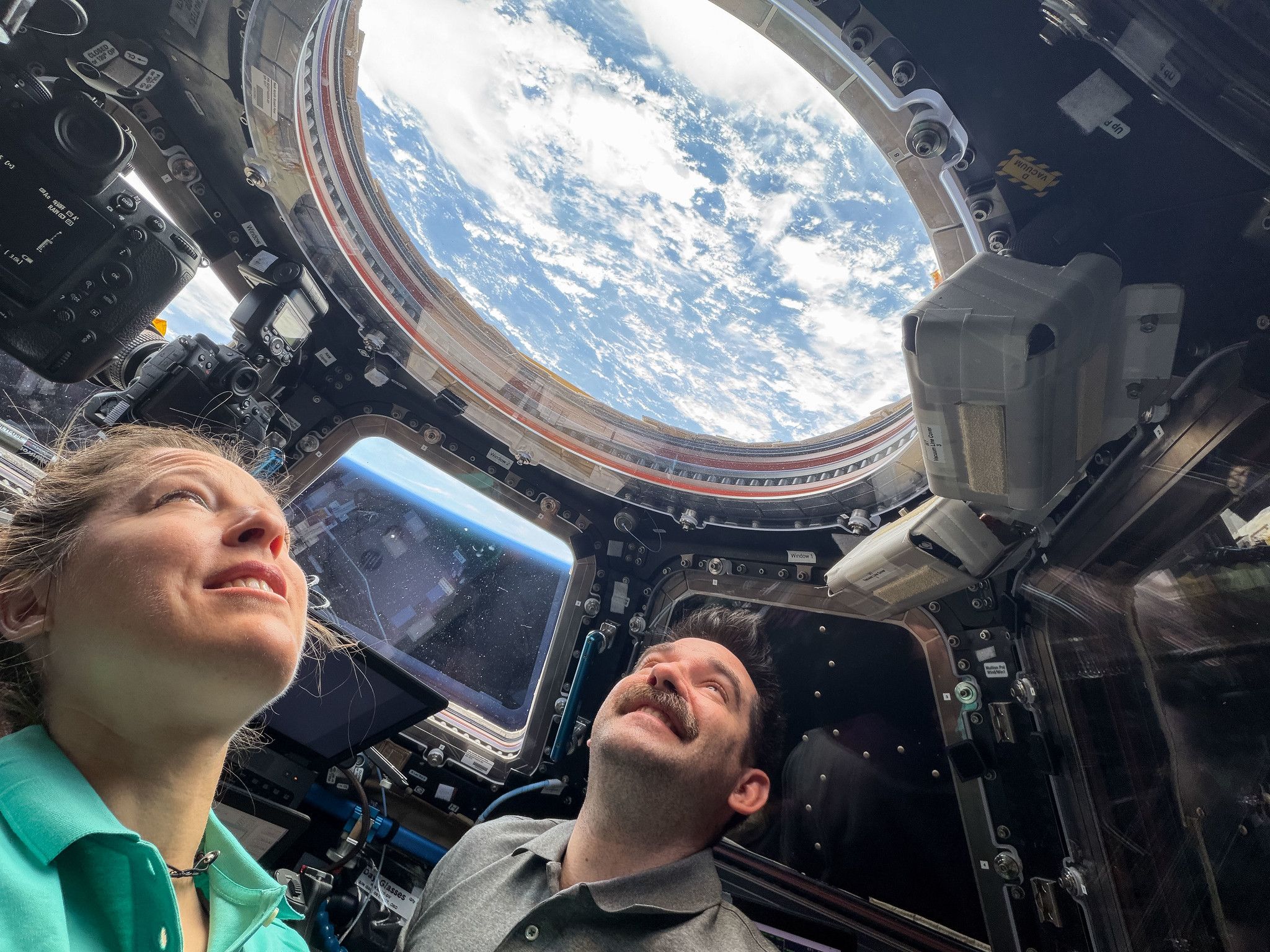 Two astronauts inside a spacecraft cockpit gaze up at a circular window showing Earth with blue sky and white clouds; surrounding panels, switches, and equipment frame the scene.